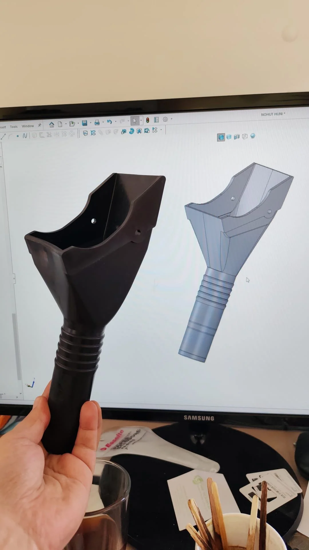 Photo of a person holding a black model of an ice cream scoop in front of a computer screen displaying a 3D design of the same scoop.