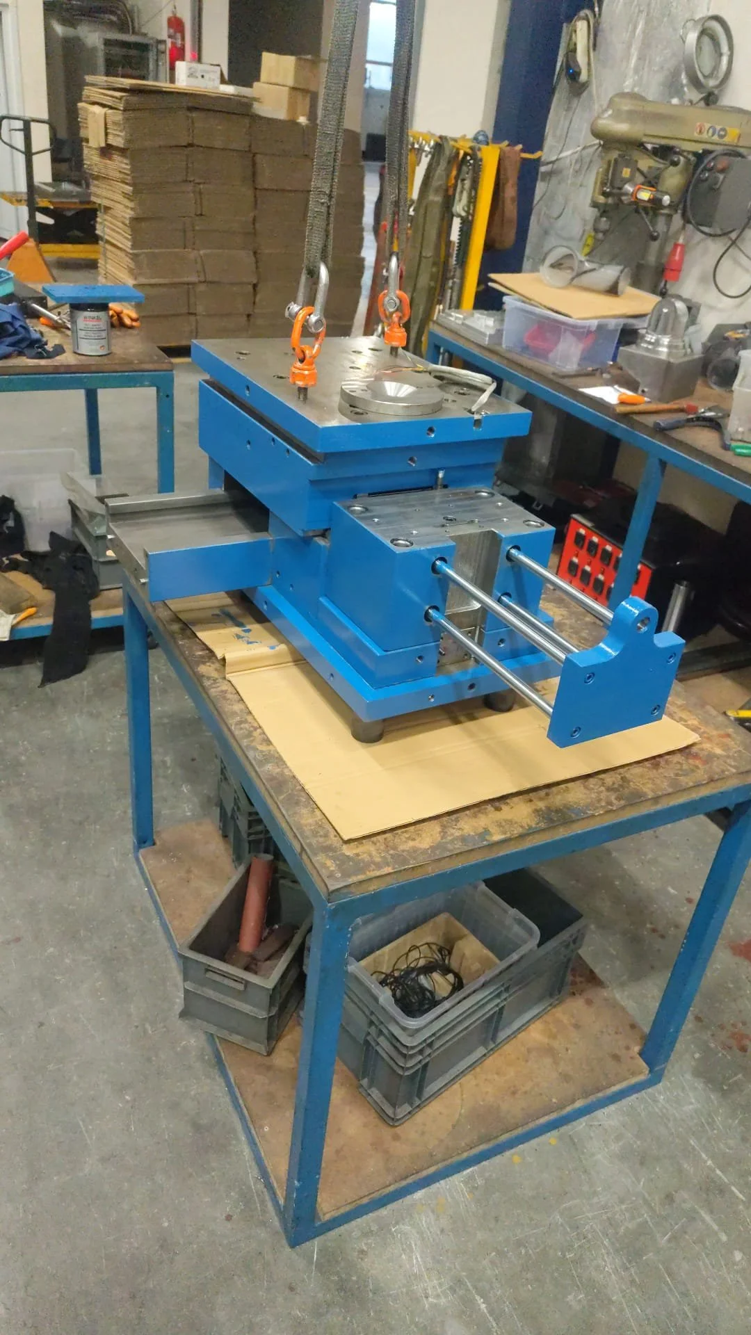 Blue mechanical workbench with metal clamp fixture and hanging straps in a workshop surrounded by tools, machine parts, and stacked cardboard boxes.