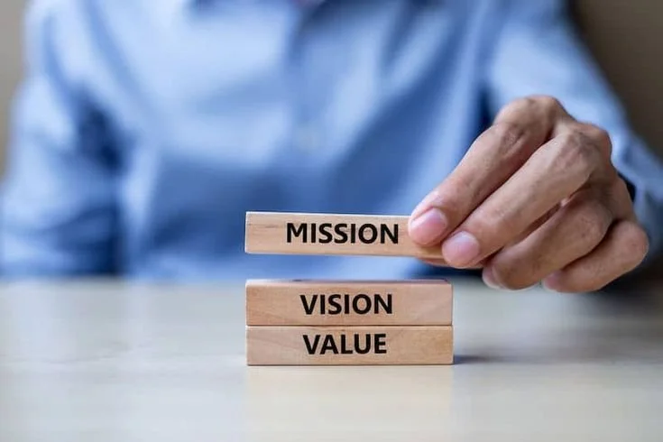 Person stacking wooden blocks labeled 'MISSION,' 'VISION,' and 'VALUE' on a table.
