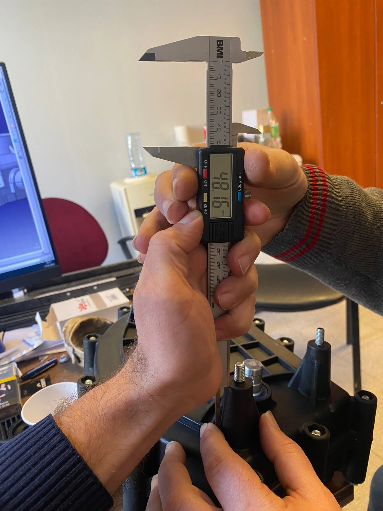 Person using a digital caliper to measure an object on a desk, with a computer monitor in the background.