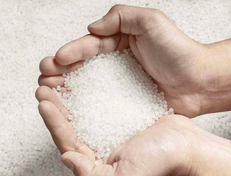 Two hands holding a handful of white granular substance, possibly rice or small beads, against a neutral background.