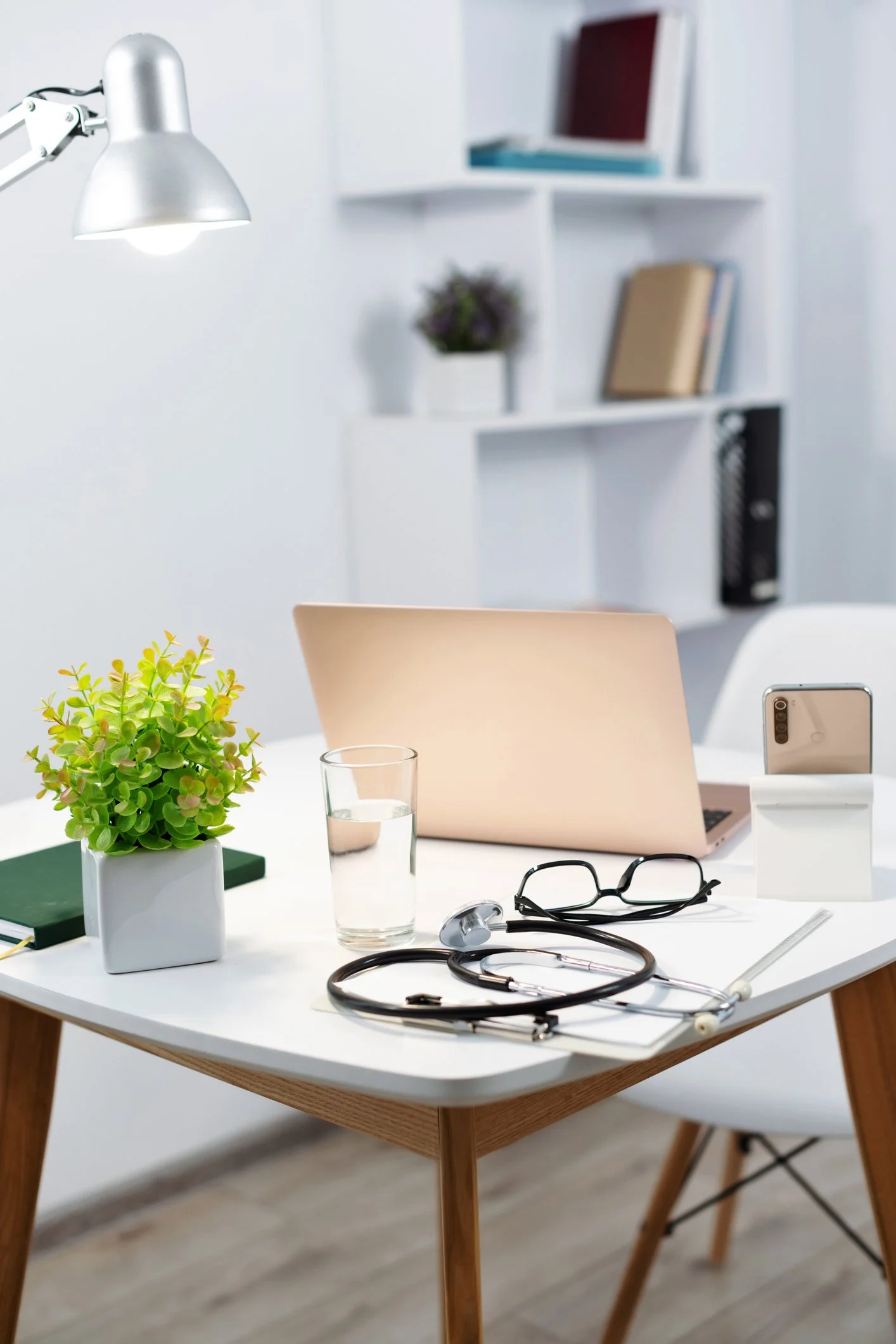 A white office desk with a plant, glass of water, glasses, stethoscope, and a laptop. There is a candle holder with a smartphone in it. In the background, a white bookshelf with books and decorations and a white chair.
