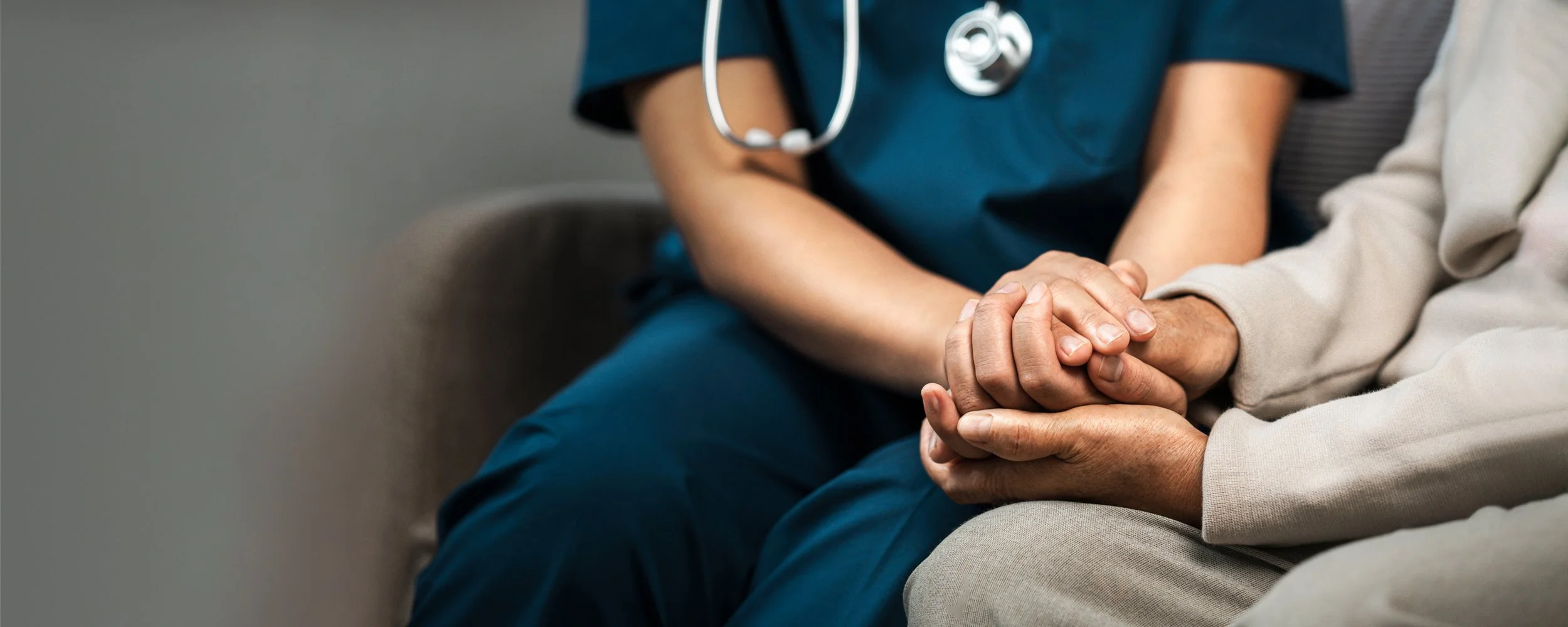 Close-up of a healthcare worker in blue scrubs with a stethoscope, holding the hand of a patient, conveying care and support.