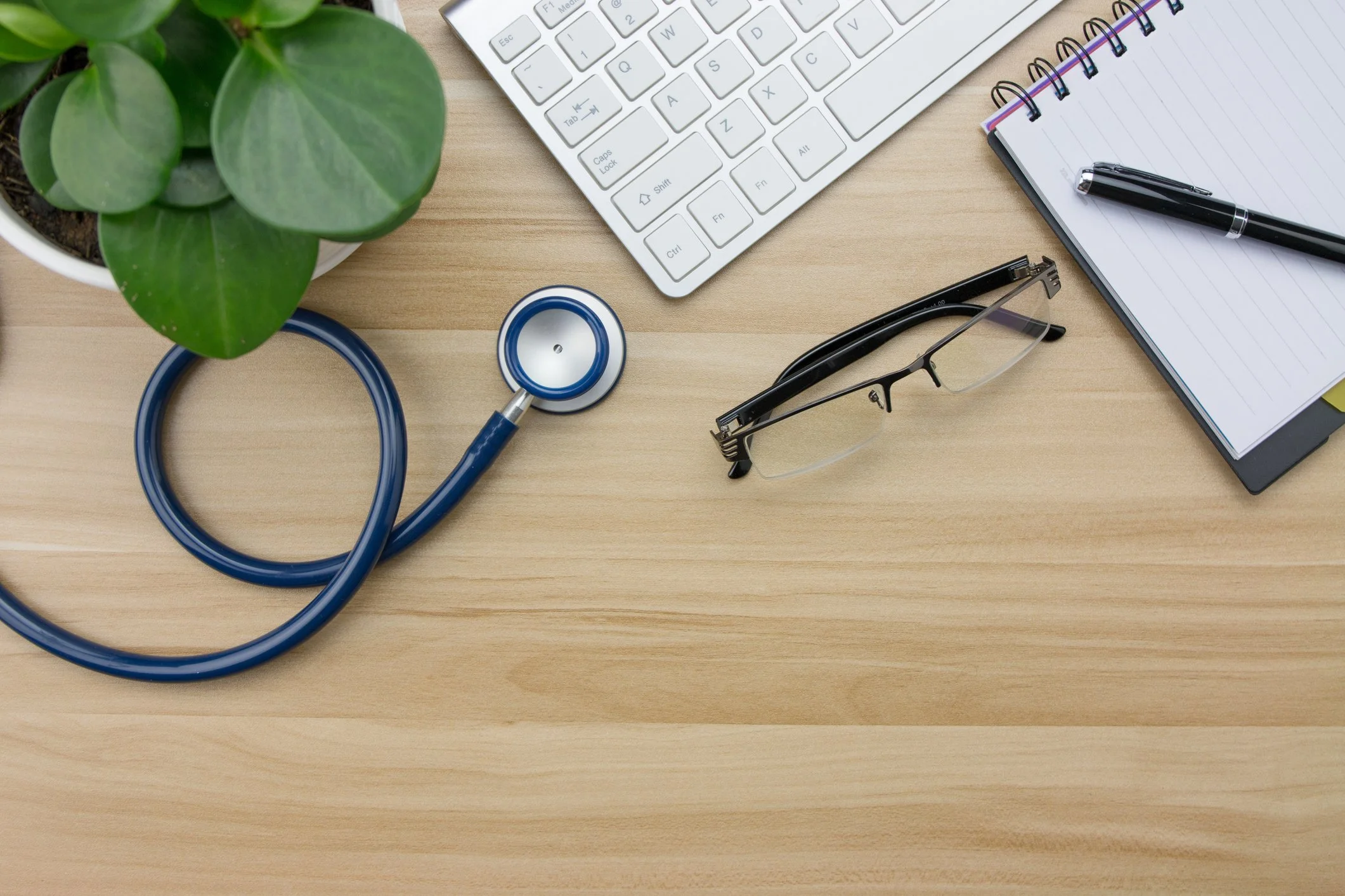 A wooden desk with a potted plant, a stethoscope, eyeglasses, a computer keyboard, a notebook with a pen, and some sticky notes.