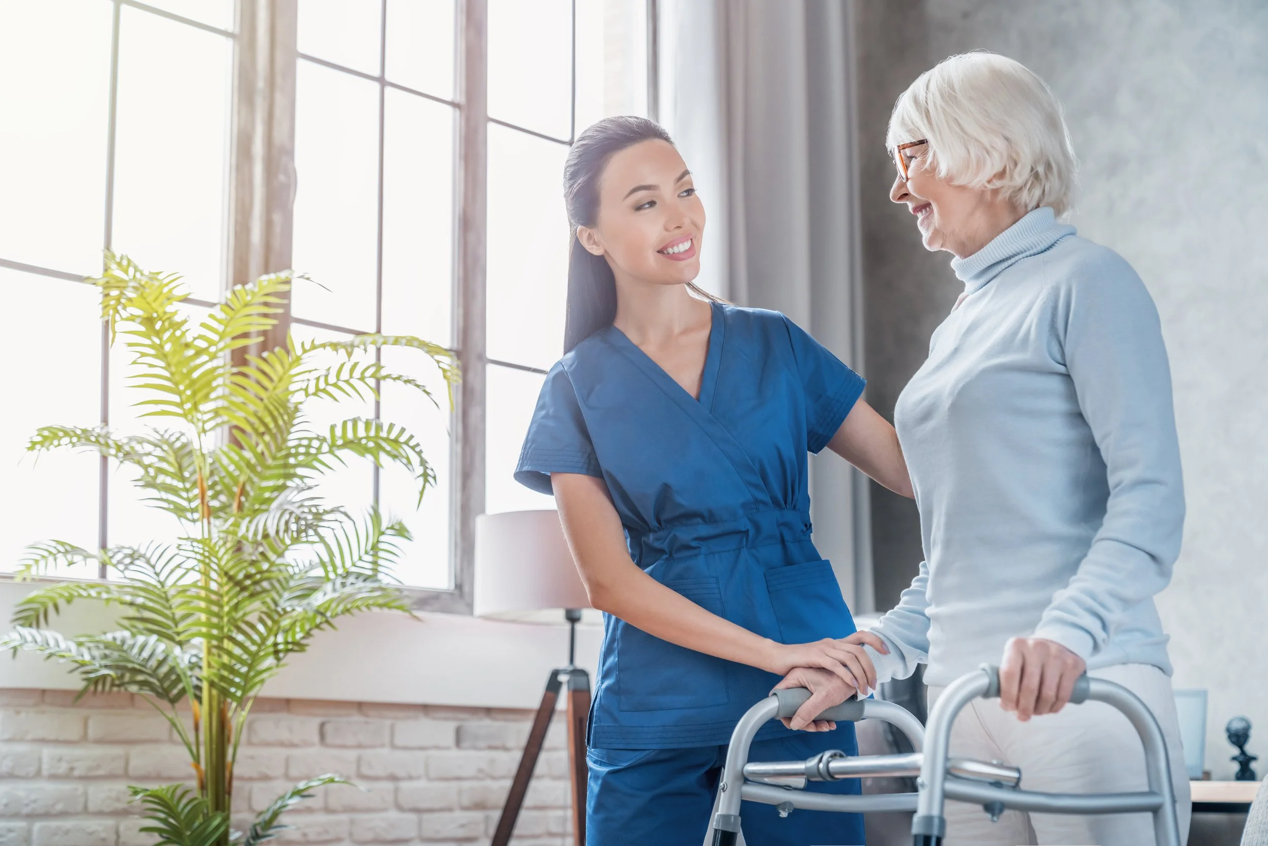 A young female nurse in blue scrubs helping an elderly woman with a walker in a brightly lit room with large windows and green plants.