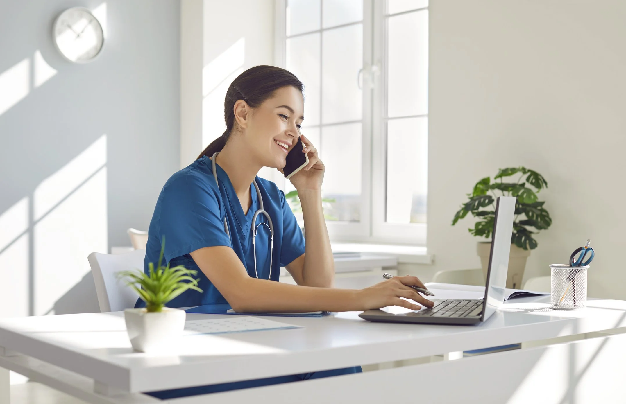 Young female healthcare professional in blue scrubs, smiling and talking on the phone while working on a laptop at a white desk.