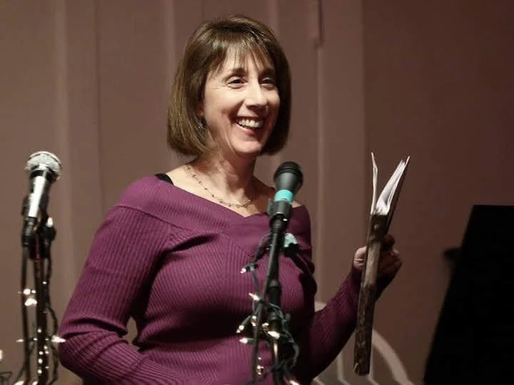 An image of Betsy Johnson from the chest up wearing a maroon sweater. She is holding a book of poetry and standing in front of two microphones, smiling.