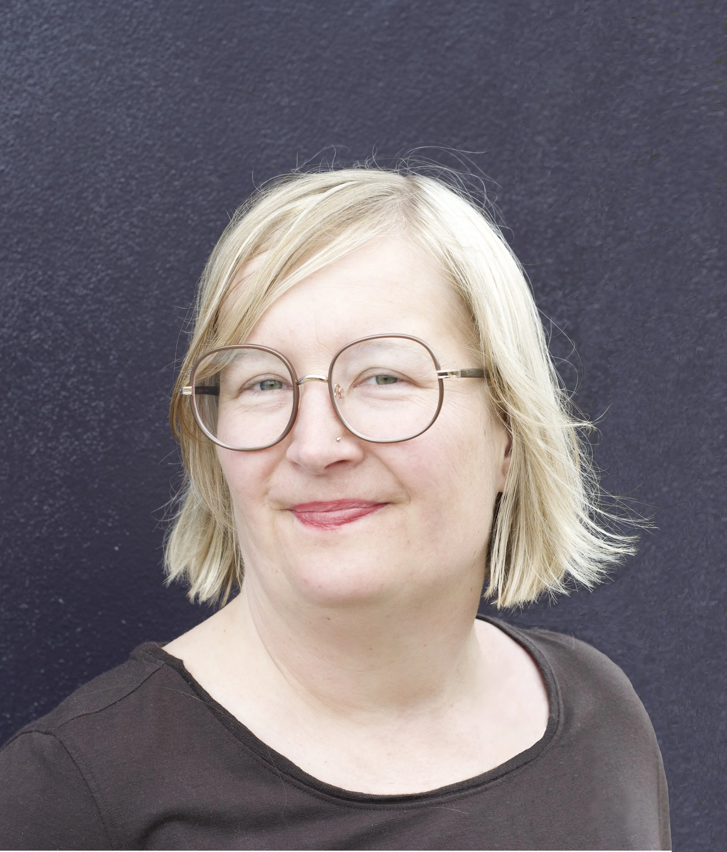 A woman with short blonde hair, glasses, and a nose piercing, wearing a black top, standing in front of a dark textured background.