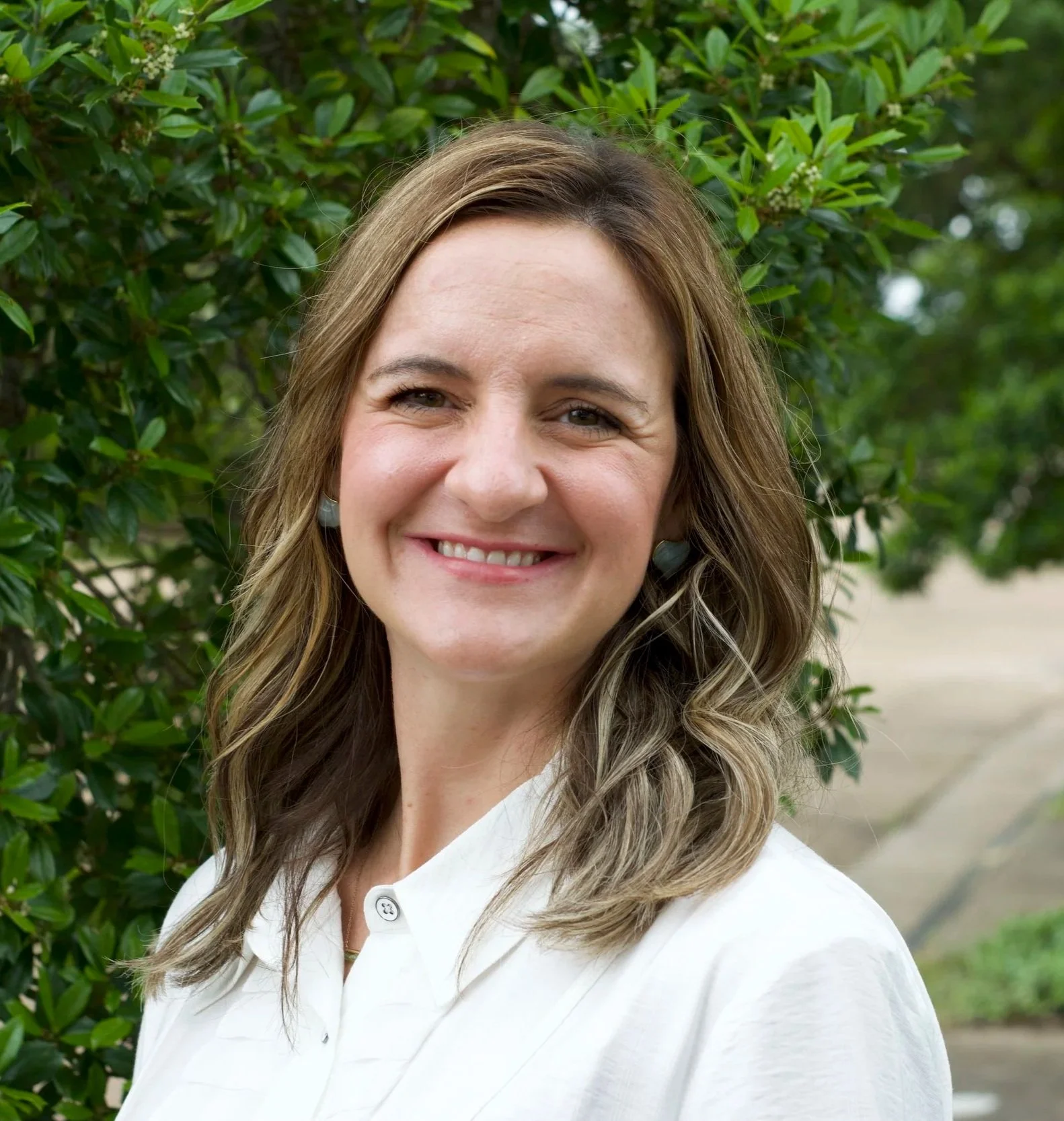 A woman with shoulder-length brown hair smiling outdoors, wearing a white shirt, standing in front of green foliage.