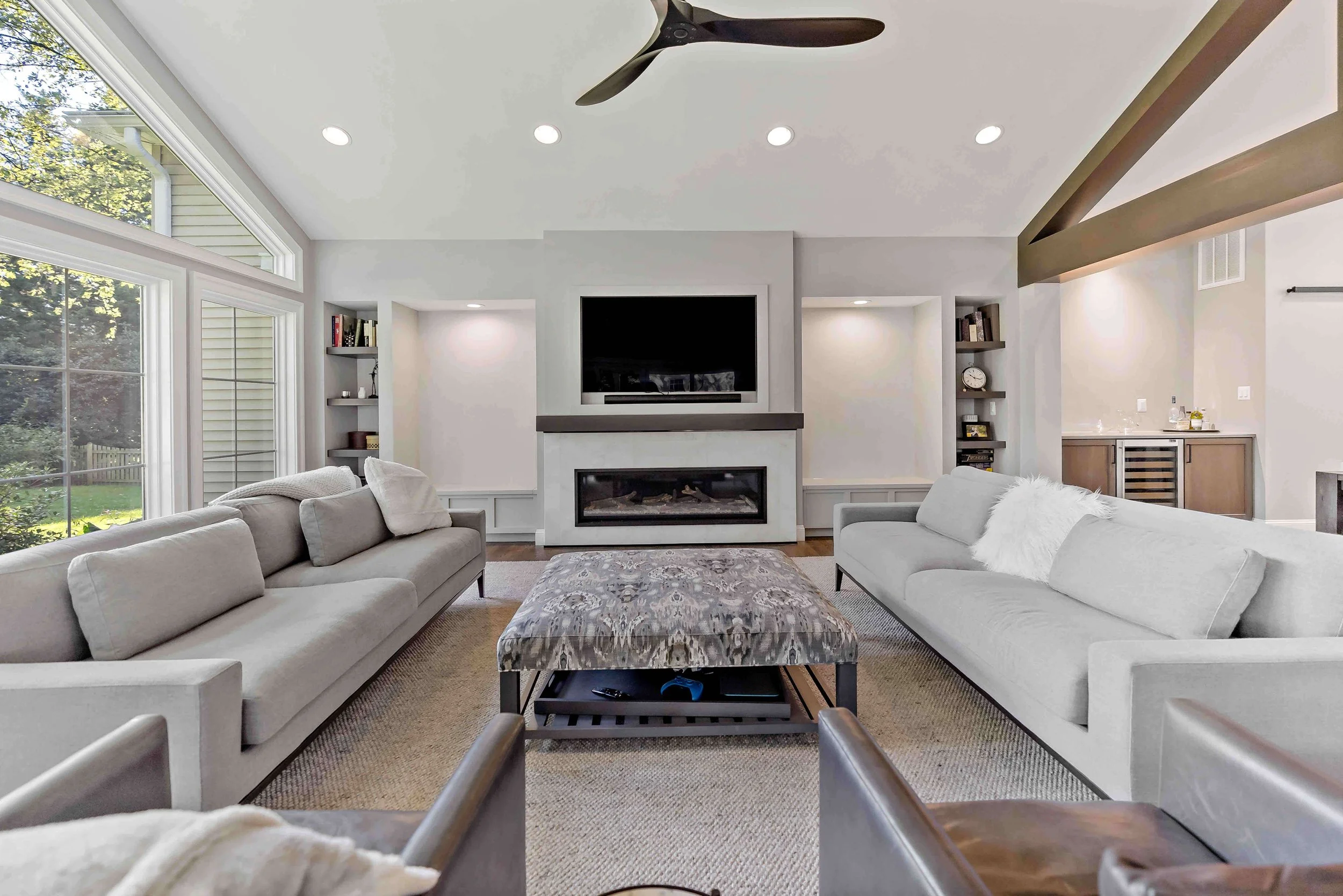 Modern living room with large windows, two light-colored sofas, a patterned ottoman, a fireplace, TV mounted above the fireplace, built-in shelves, and visible wooden beams on the ceiling.