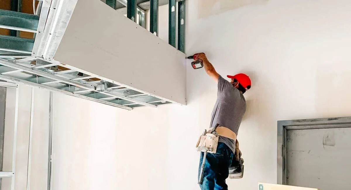 Construction worker installing drywall on a ceiling with a power drill.