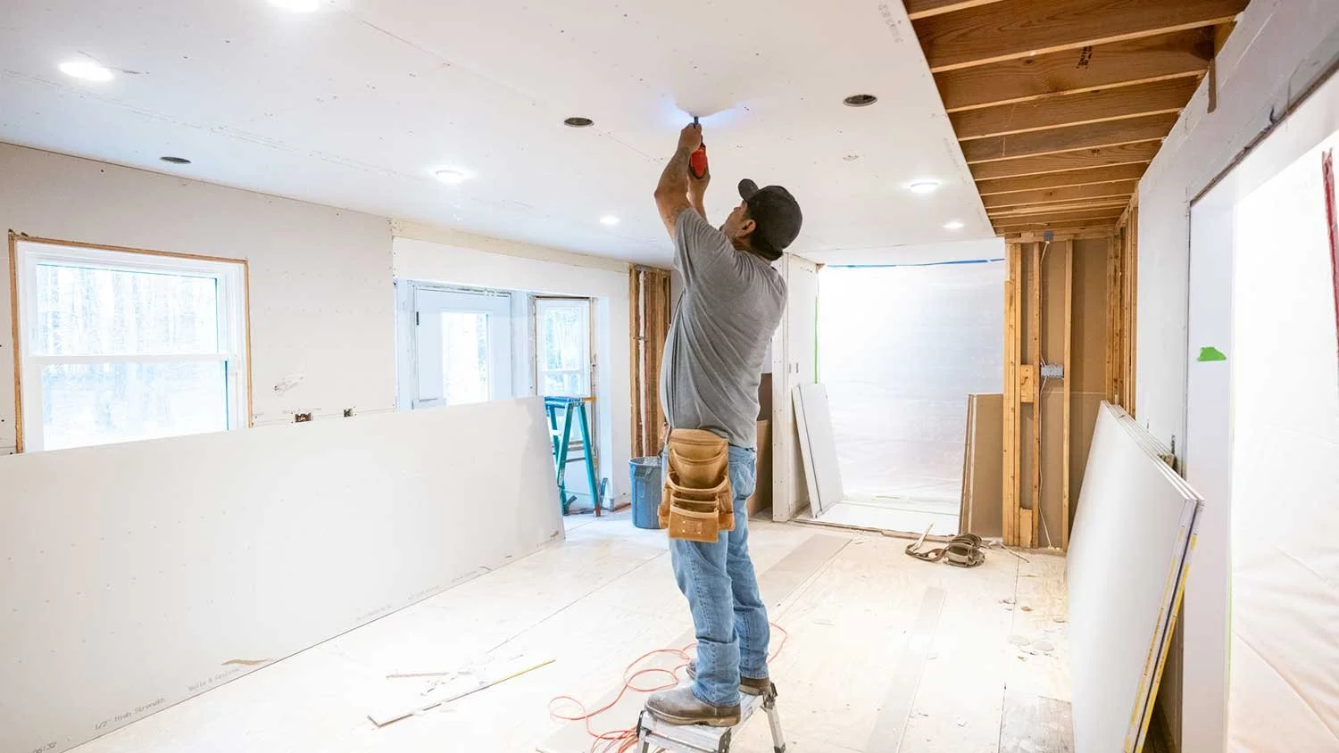 A construction worker standing on a small step ladder, installing a ceiling light fixture in a room under renovation.