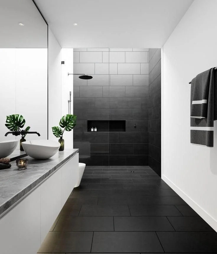 Modern bathroom with black tiled walk-in shower, white walls, gray countertop with two vessel sinks, and a large mirror. Towel hanging on the right side and decorative plants on the countertop.