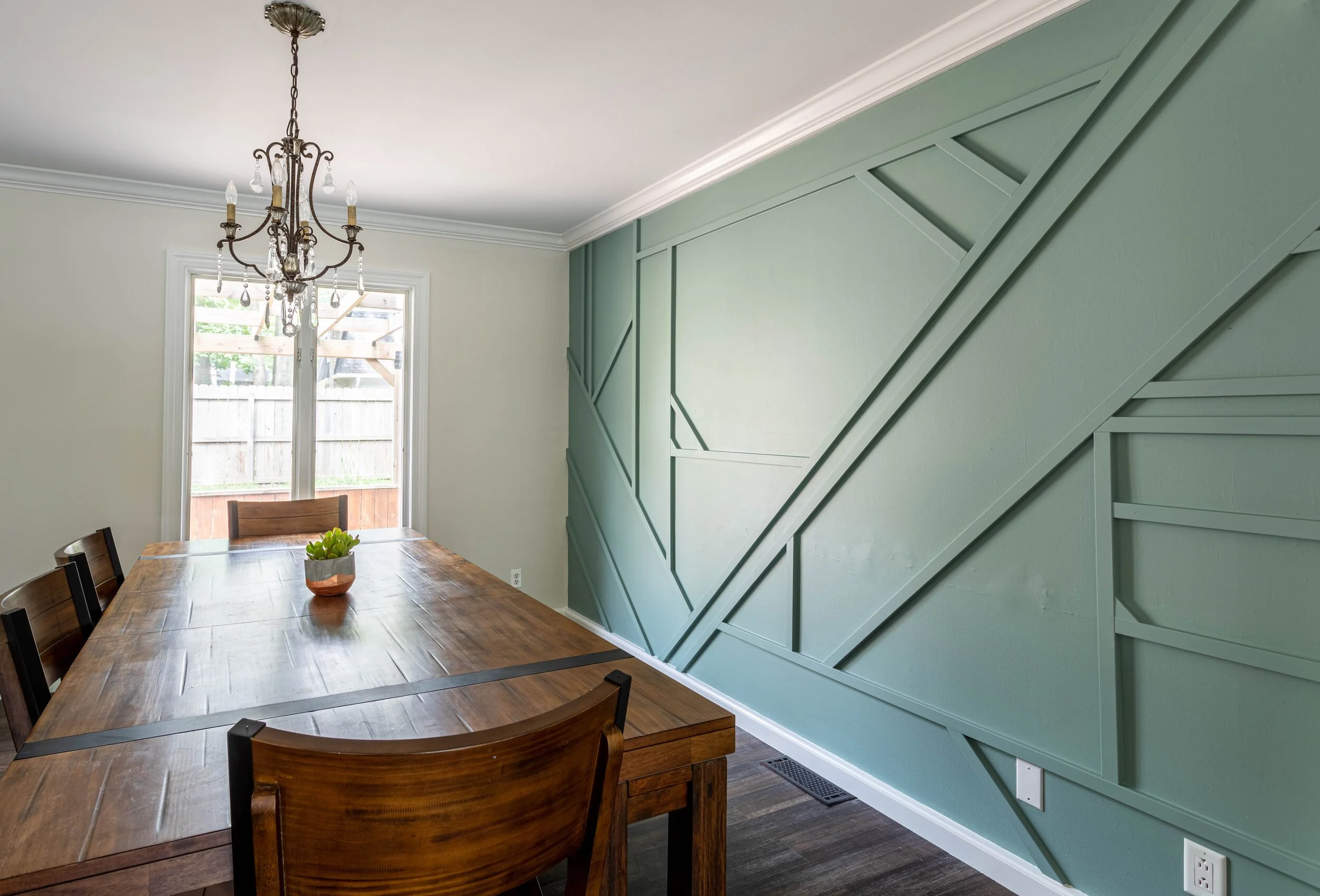Dining room with wooden table, seven chairs, green geometric-patterned accent wall, chandelier, and a window showing a fenced backyard.