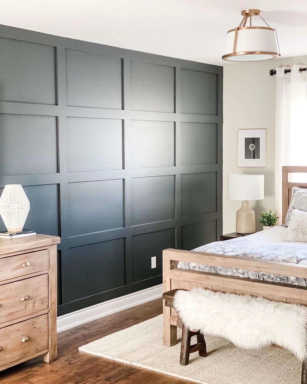 A bedroom featuring a dark gray paneled accent wall, light wood furniture including a dress and bench with a faux fur throw, a nightstand with a lamp, a framed black and white flower art, and a window with white curtains.