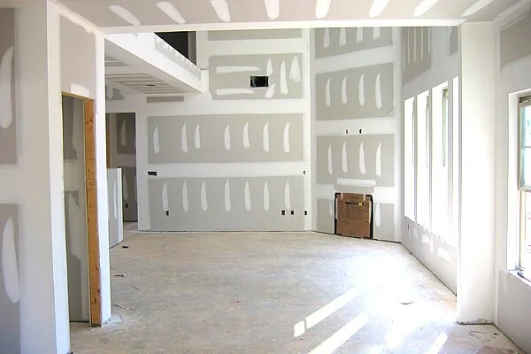 An empty room under construction with drywall panels installed on the walls and ceiling, with unfinished flooring and multiple windows letting in natural light.