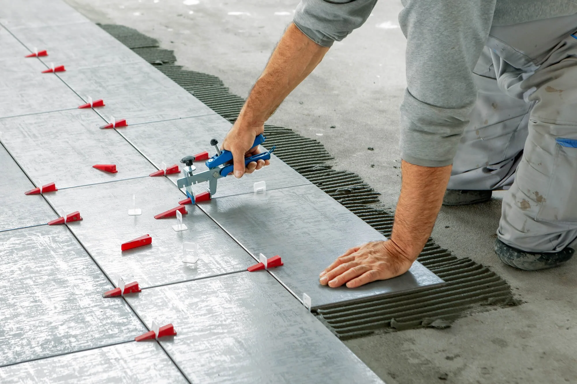 A worker kneels on a concrete floor, installing large grey tiles with the help of tile spacers and a tile leveling system.