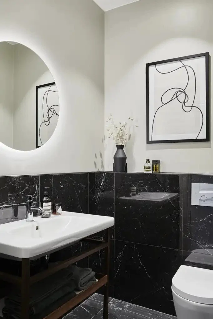 Modern bathroom with black marble lower walls, white sink, decorative abstract black line art on the wall, a round mirror, a black vase with white flowers, and framed abstract artwork.