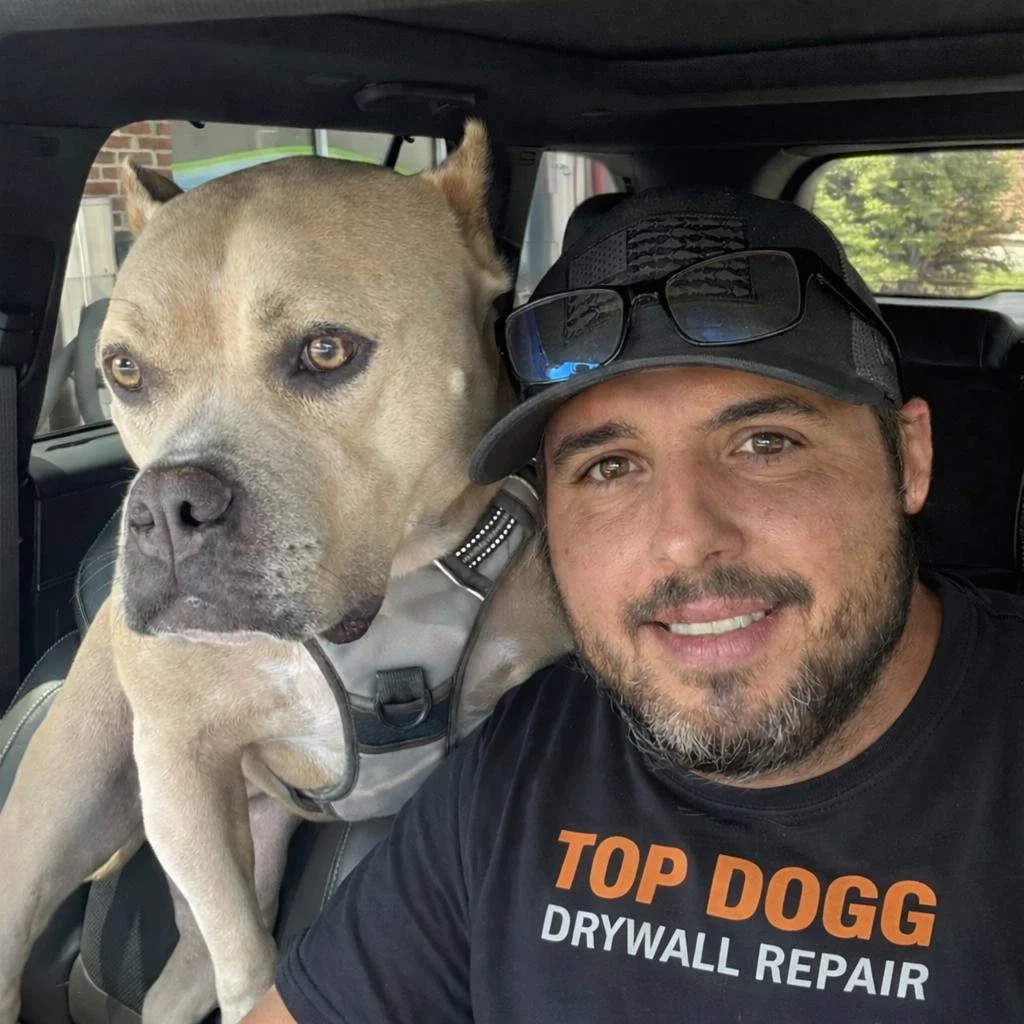 A smiling man wearing a black cap and a black T-shirt with the words 'TOP DOGG DRYWALL REPAIR' is sitting next to a large dog inside a vehicle. The dog has a tan coat, black markings around its eyes, and is wearing a harness.
