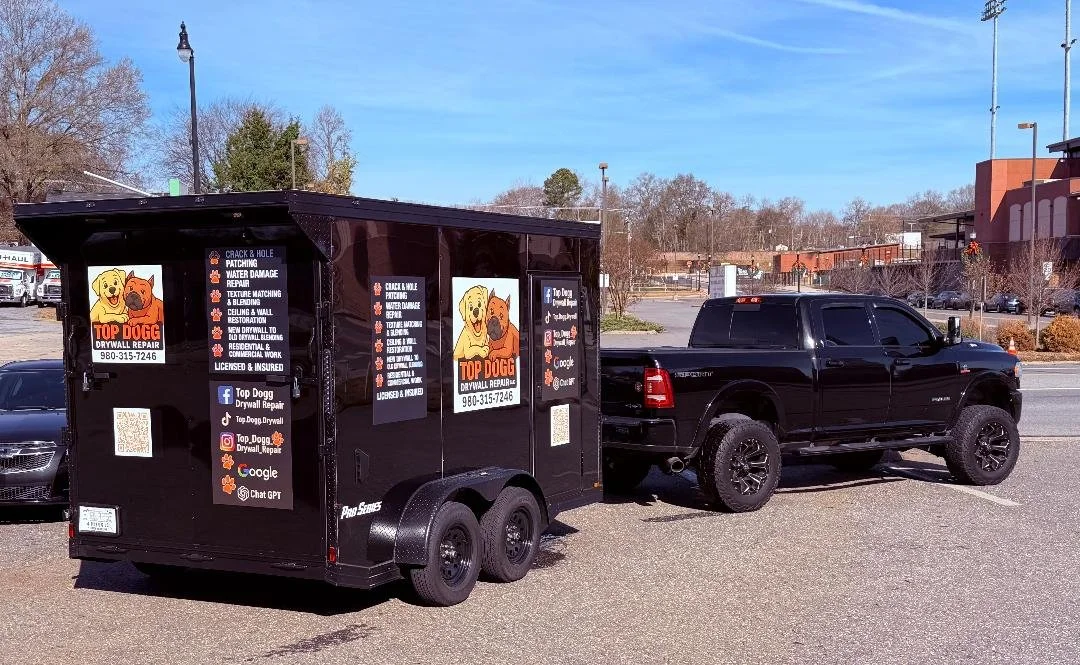 A black trailer with advertisements for Top Dogg Drywall Repair, parked in a parking lot with a black pickup truck attached. The trailer displays company services, icons for social media platforms and contact information, including a phone number and Facebook, Instagram, and Google icons.