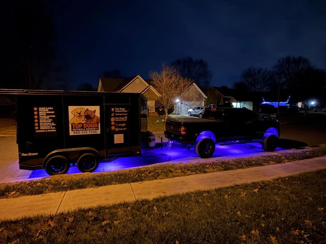 A black truck parked on a residential street at night, attached to a black trailer with blue underglow lights. The trailer advertises Top Dog Drywall Repair with a logo of a smiling dog and a dog with a sad face, along with contact information and social media icons.