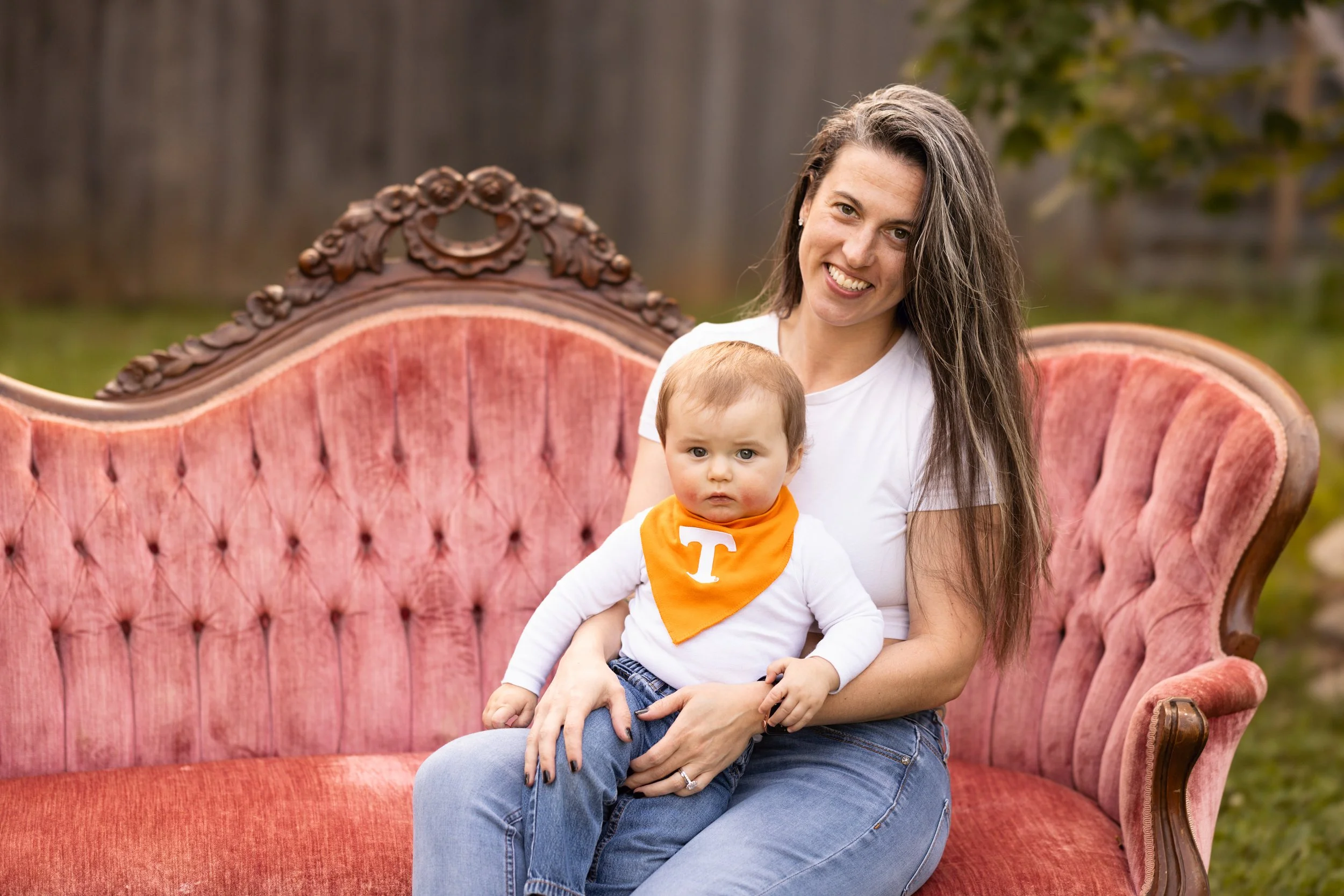 A woman and a young boy sitting on a vintage pink velvet sofa outdoors with a wooden fence and greenery in the background.