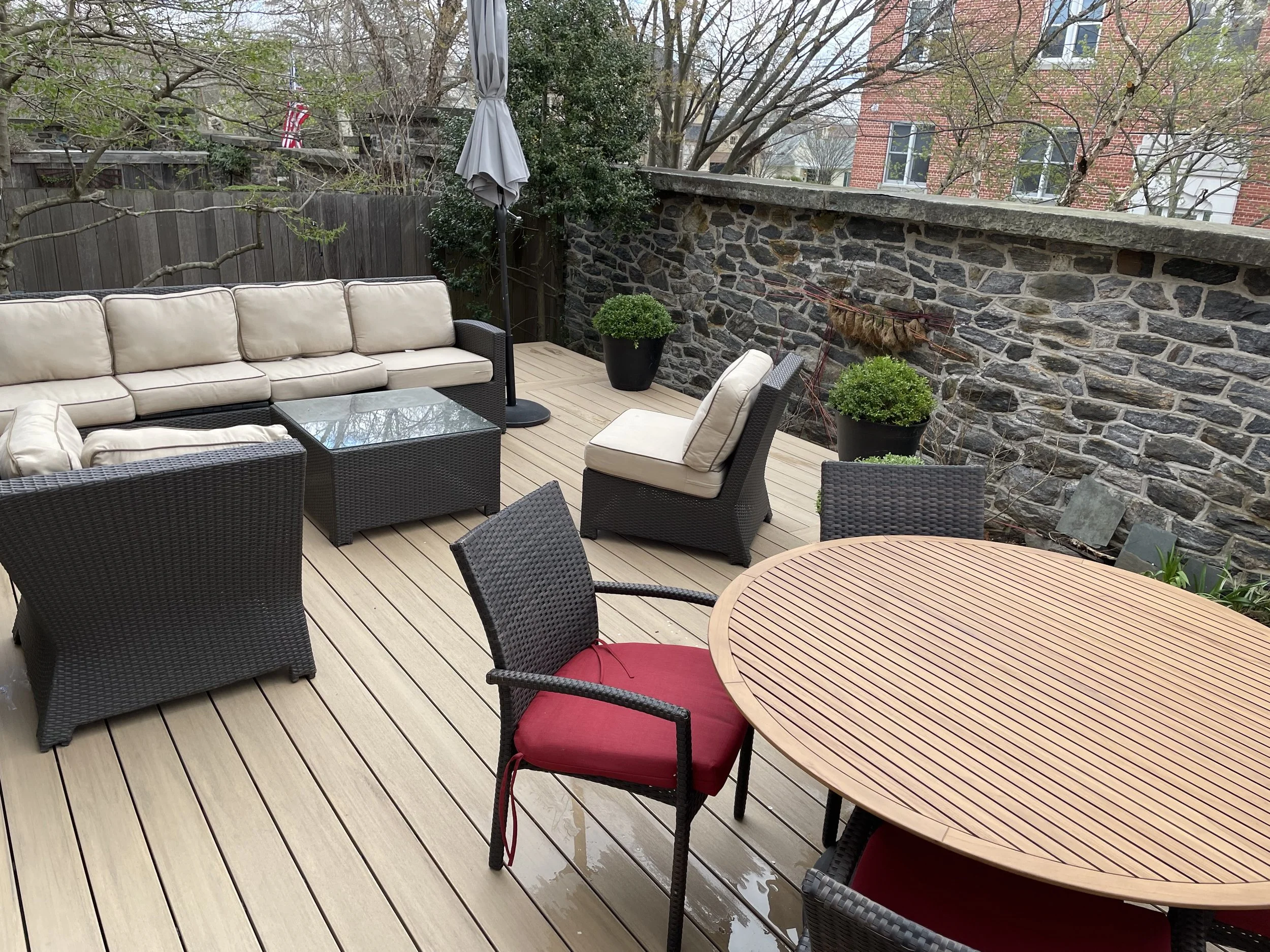 An outdoor patio with woven furniture, a beige cushioned sectional sofa, a round wooden dining table with red cushions, a glass coffee table, and potted plants, enclosed by a stone wall and wooden fence.