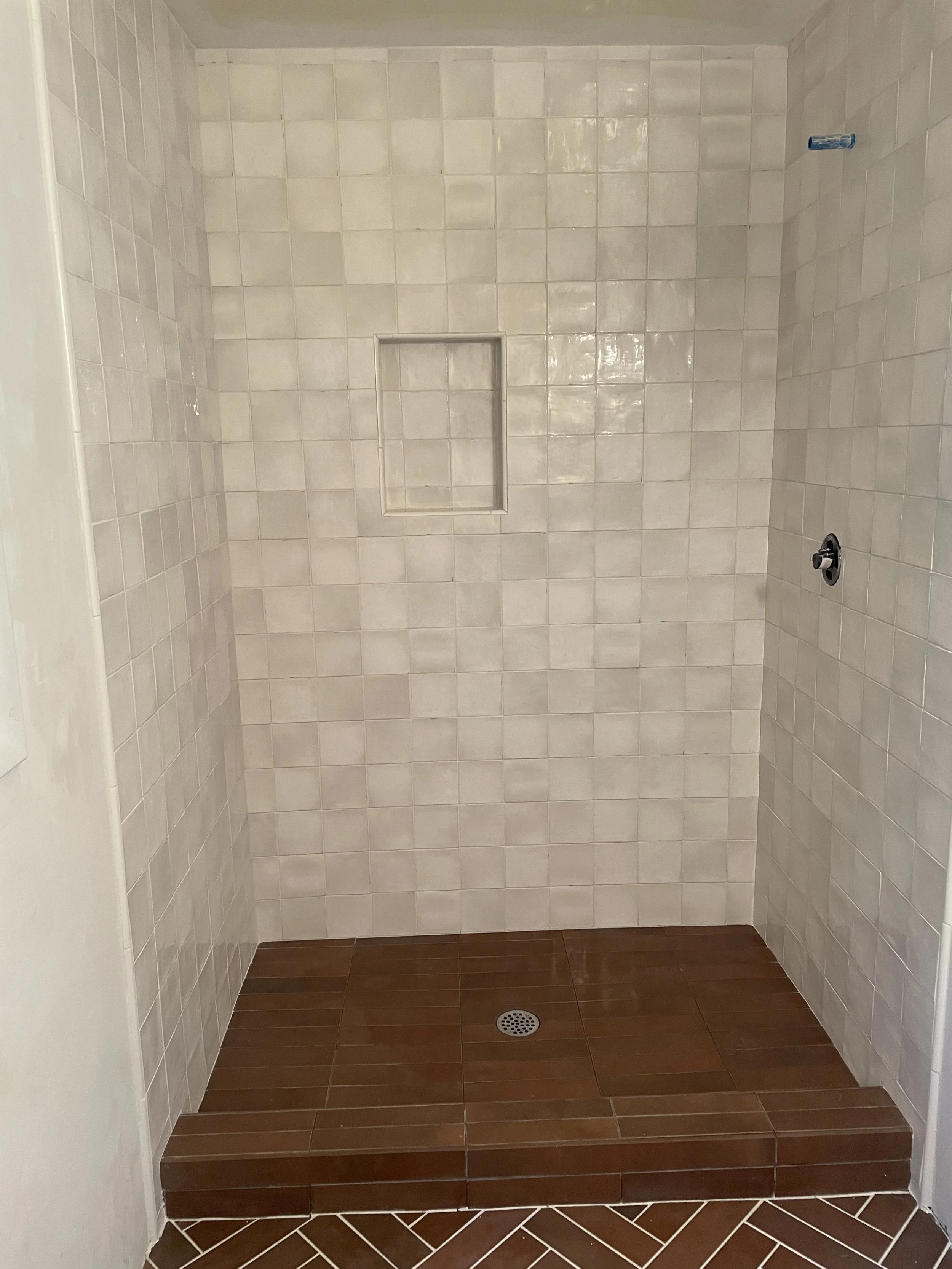 Empty walk-in shower with white tiled walls, built-in niche, brown tiled floor, and a floor drain.