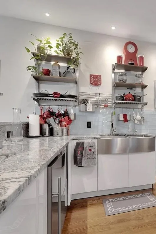 Kitchen with white cabinets, granite countertop, stainless steel sink, open shelving with red kitchenware, P-trap dish drying rack, and decorative red clock and accessories.