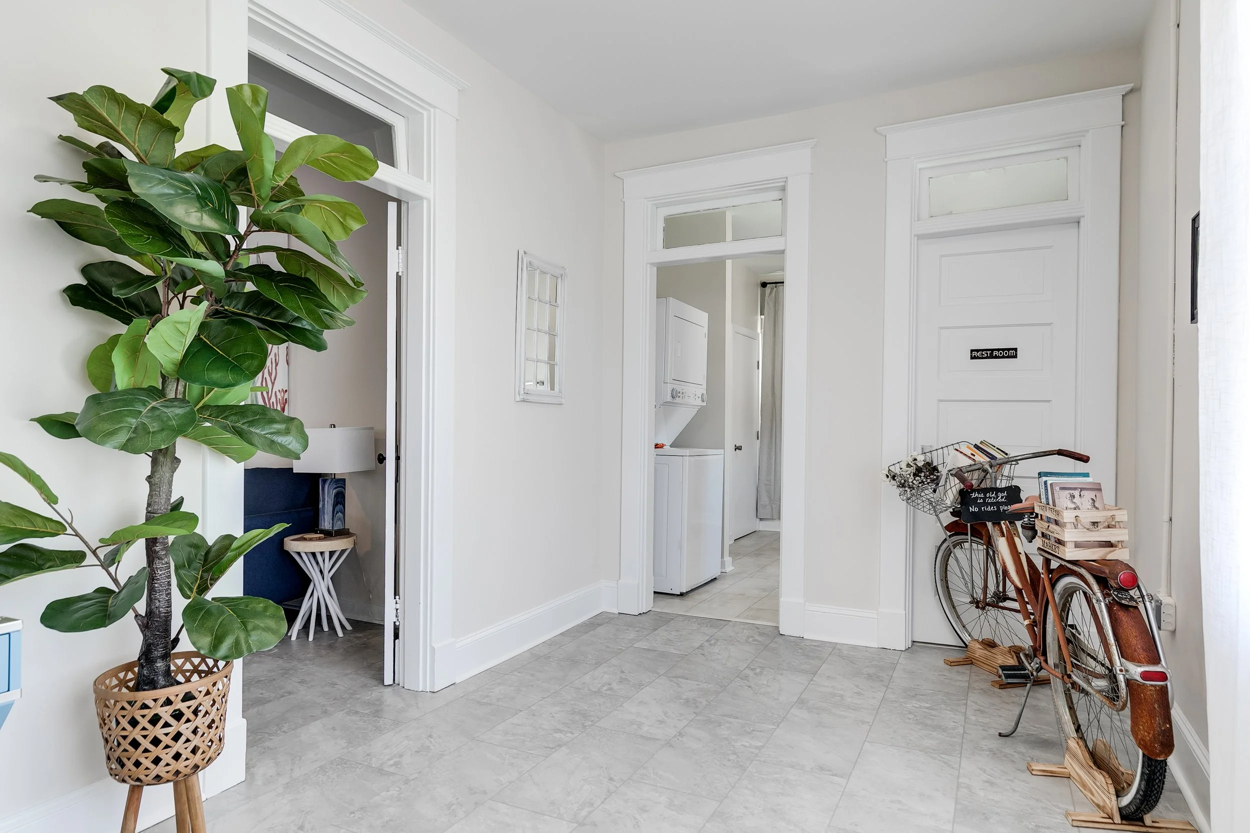 Entryway with a large potted plant on a stand, a vintage bicycle with baskets near a door labeled 'Rest Room', and a laundry area with a stacked washer and dryer.