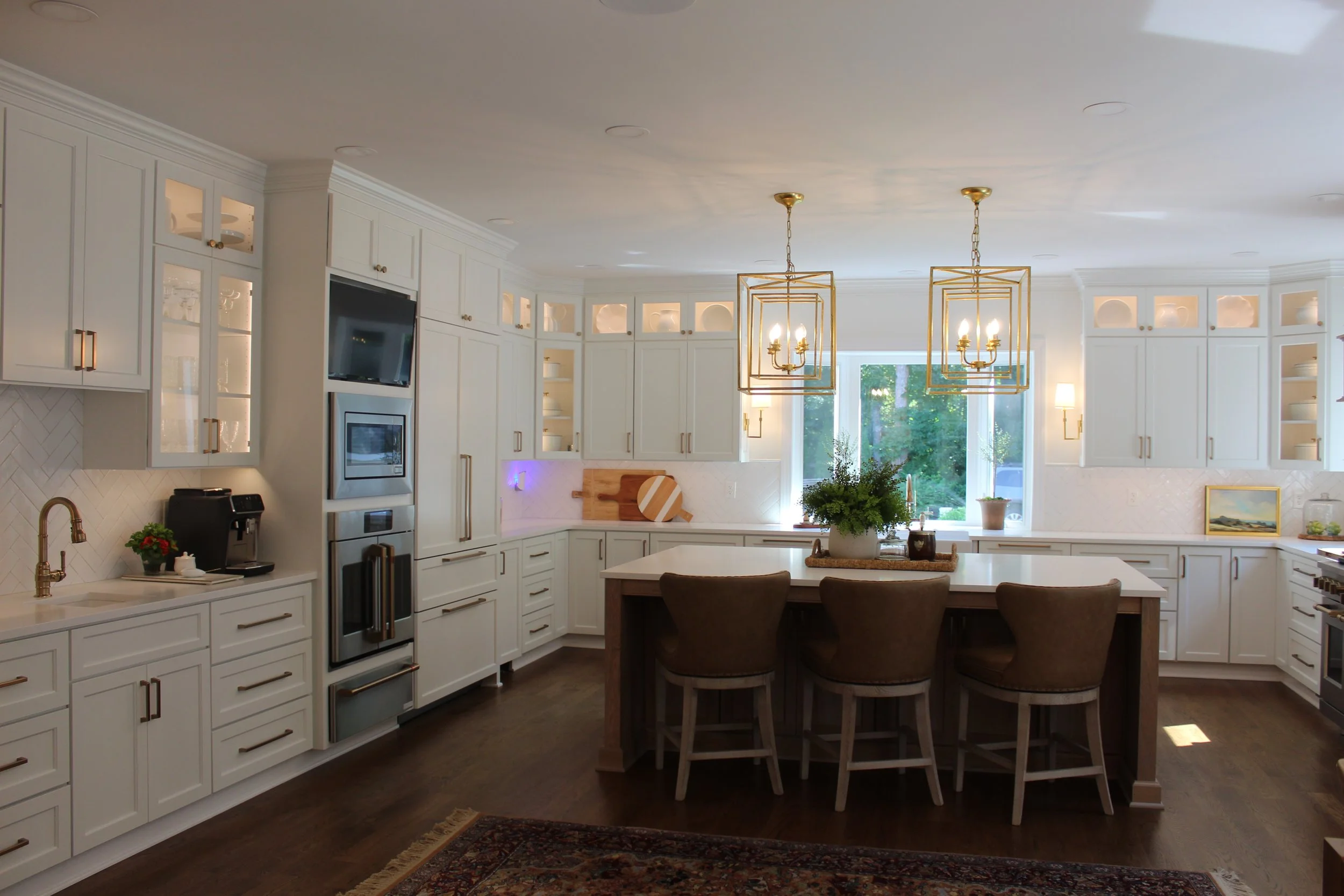 Bright white kitchen with a central island, three chairs, gold chandeliers, and large window overlooking greenery.