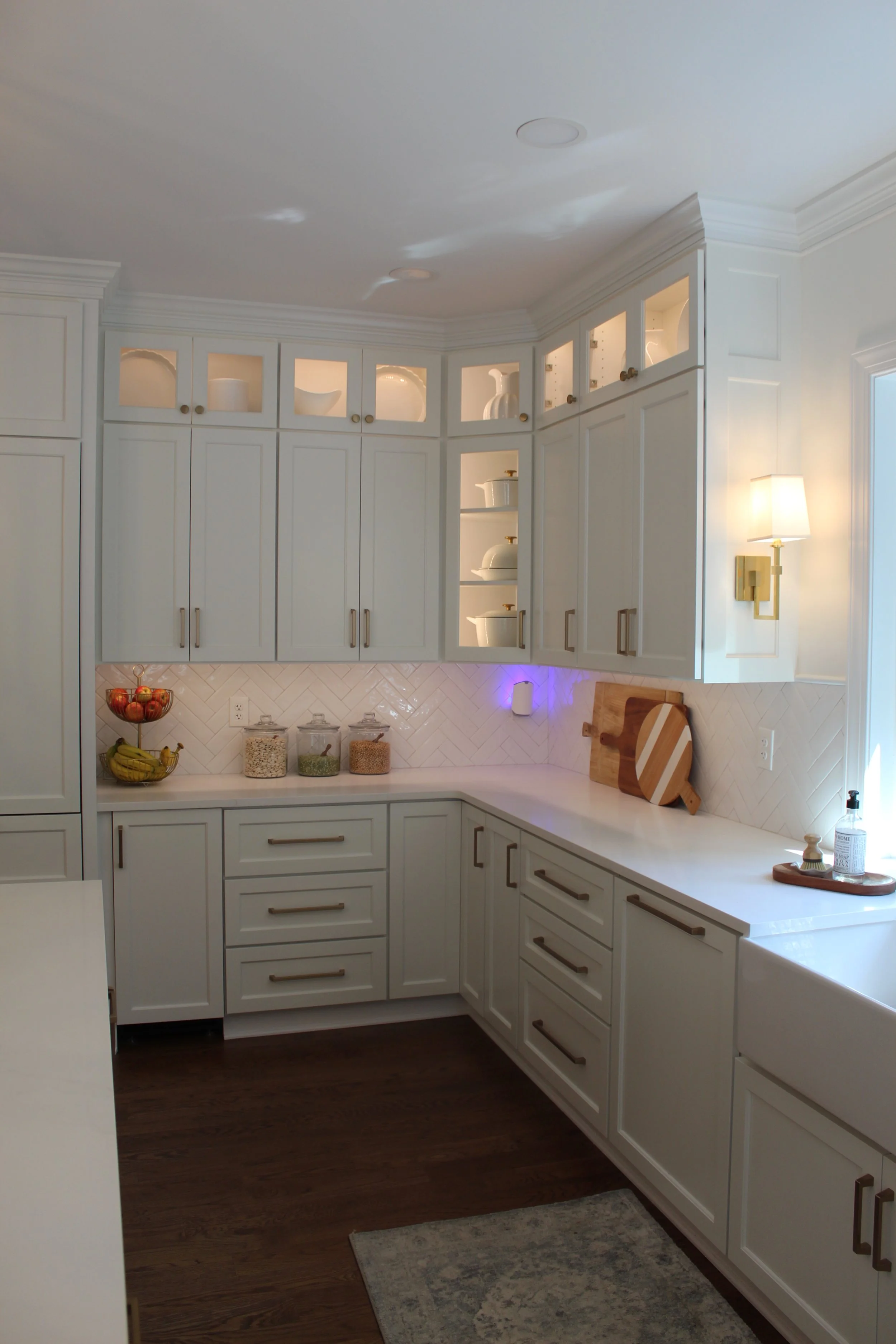 Bright white L-shaped kitchen with upper glass-front cabinets filled with white dishes, lower cabinets with modern metal handles, a dark hardwood floor, and a white countertop with fruit bowl, jars of grains, cutting boards, and soap dispenser.