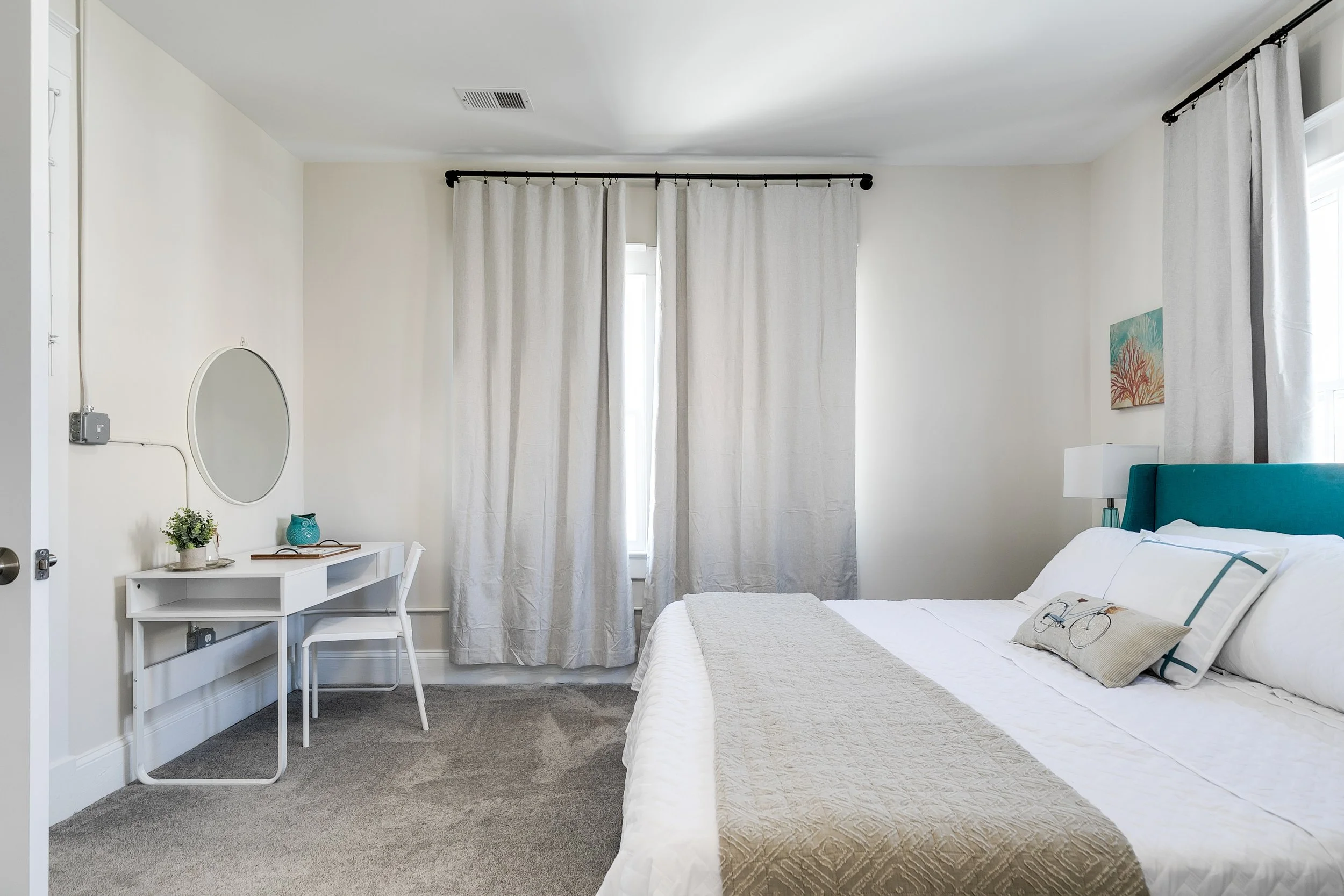 A bedroom with a white bed and teal headboard, white curtains, a small white desk with a mirror and decorative objects, beige carpet, and a framed sea-themed artwork on the wall.
