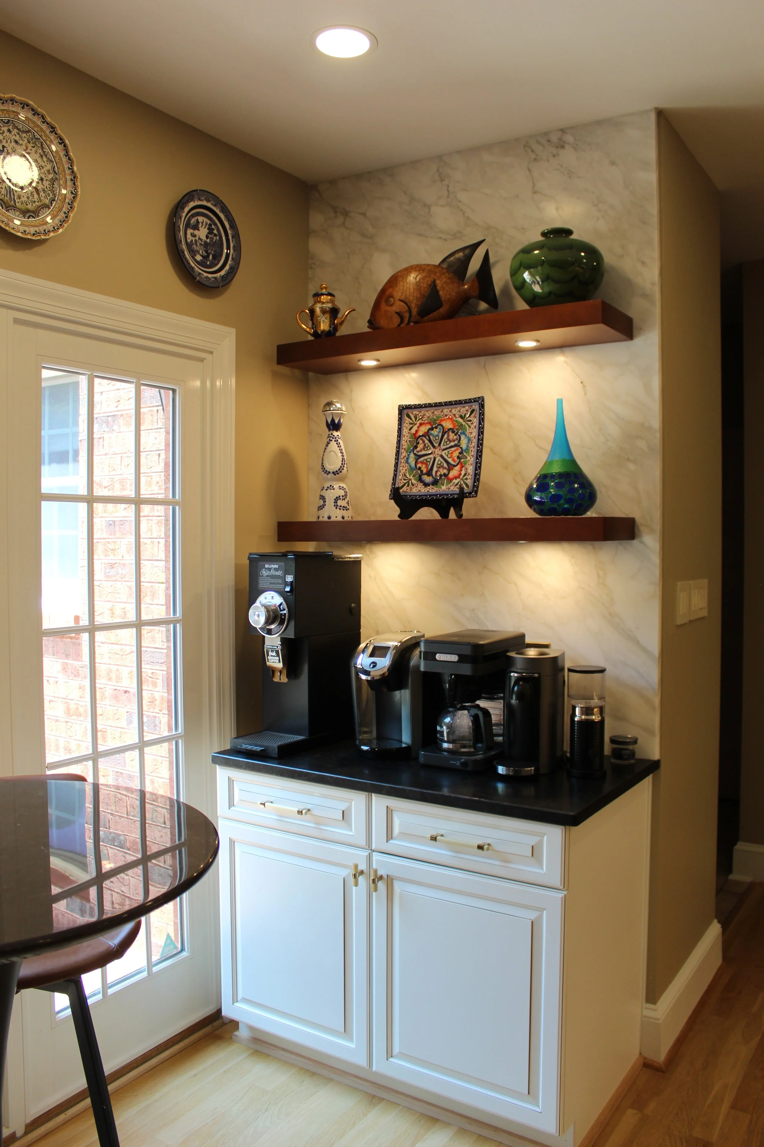 Coffee station with two wooden shelves displaying decorative vases, plates, and a fish sculpture, next to a window with a sliding glass door, and coffee machines on a white cabinet.