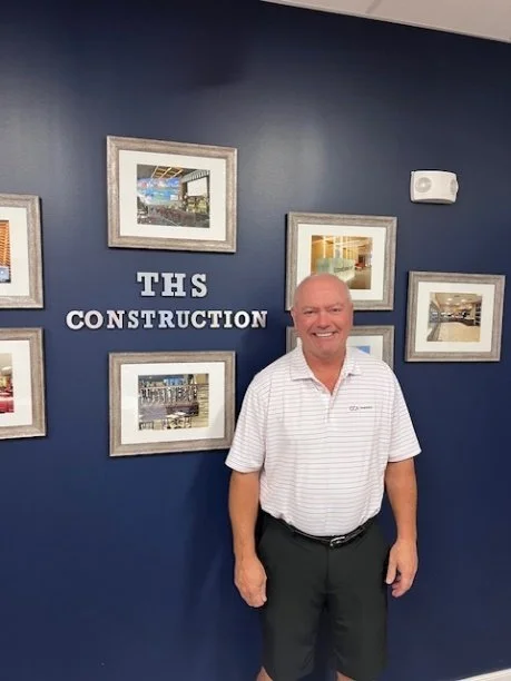 A man smiling in front of a dark blue wall with framed pictures and a sign that says "THS CONSTRUCTION."
