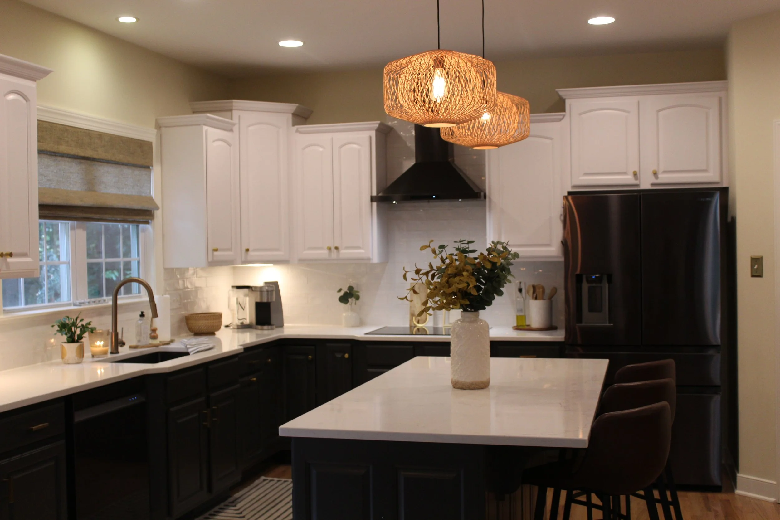 Modern kitchen with white upper cabinets, black lower cabinets, kitchen island, black refrigerator, wall-mounted oven, coffee maker, potted plants, and decorative light fixtures.