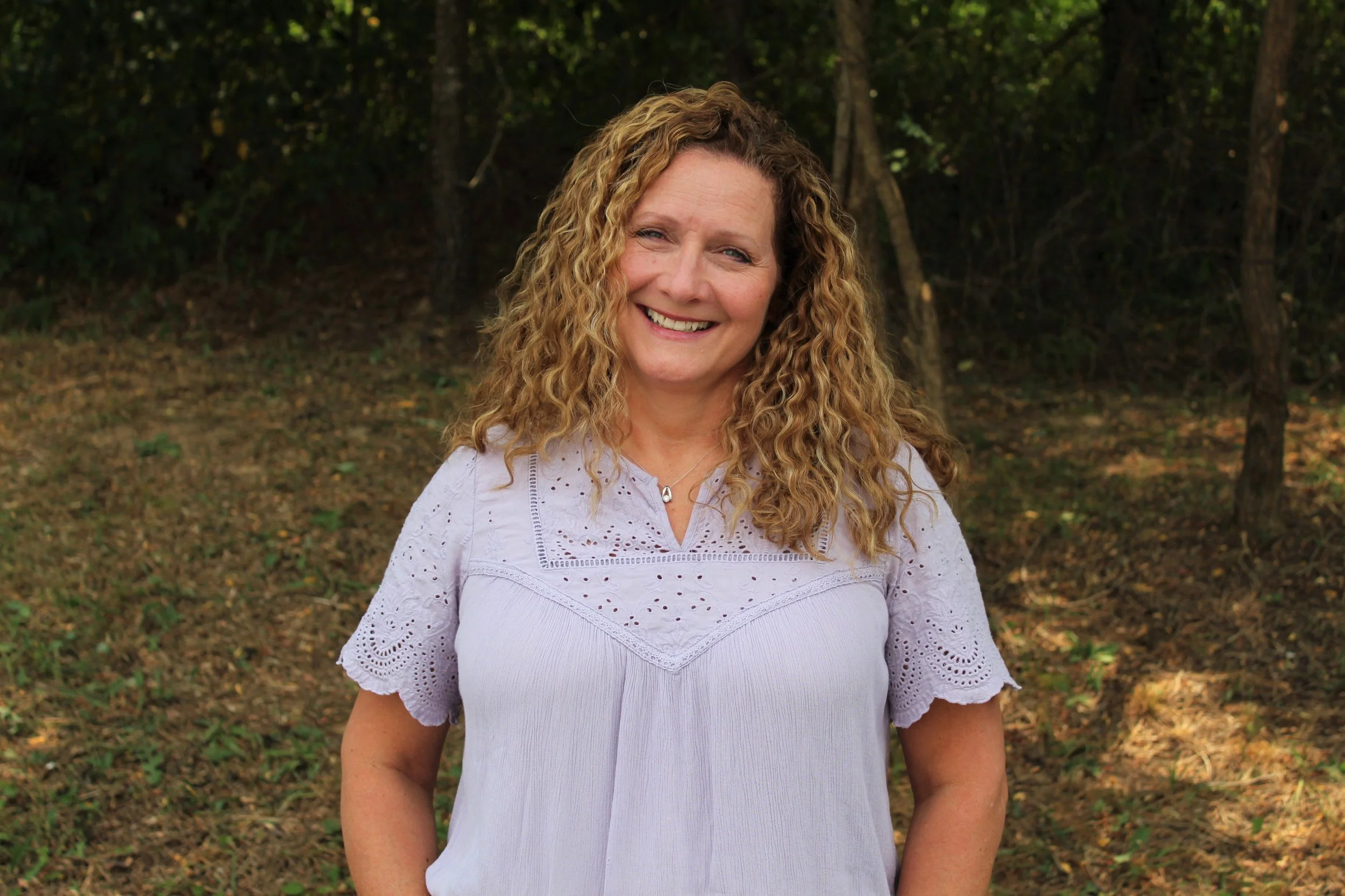 A smiling middle-aged woman with long, curly red hair standing outdoors in front of a wooded area.