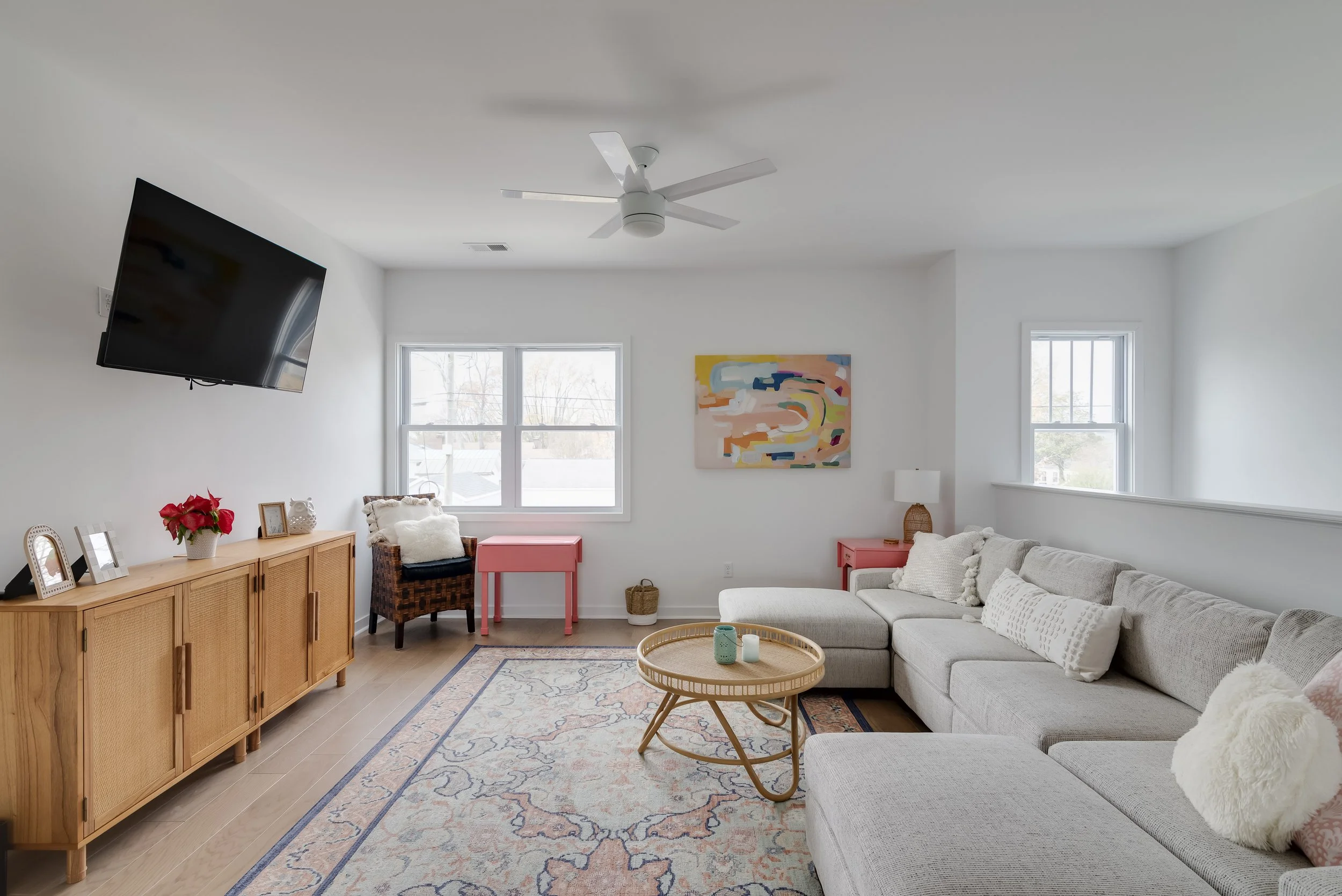 Living room with a beige sectional sofa, a wooden sideboard with picture frames and a poinsettia plant, a pink side table, a wicker chair with pillows, a colorful abstract wall art, a round wooden coffee table with candles, a patterned area rug, and