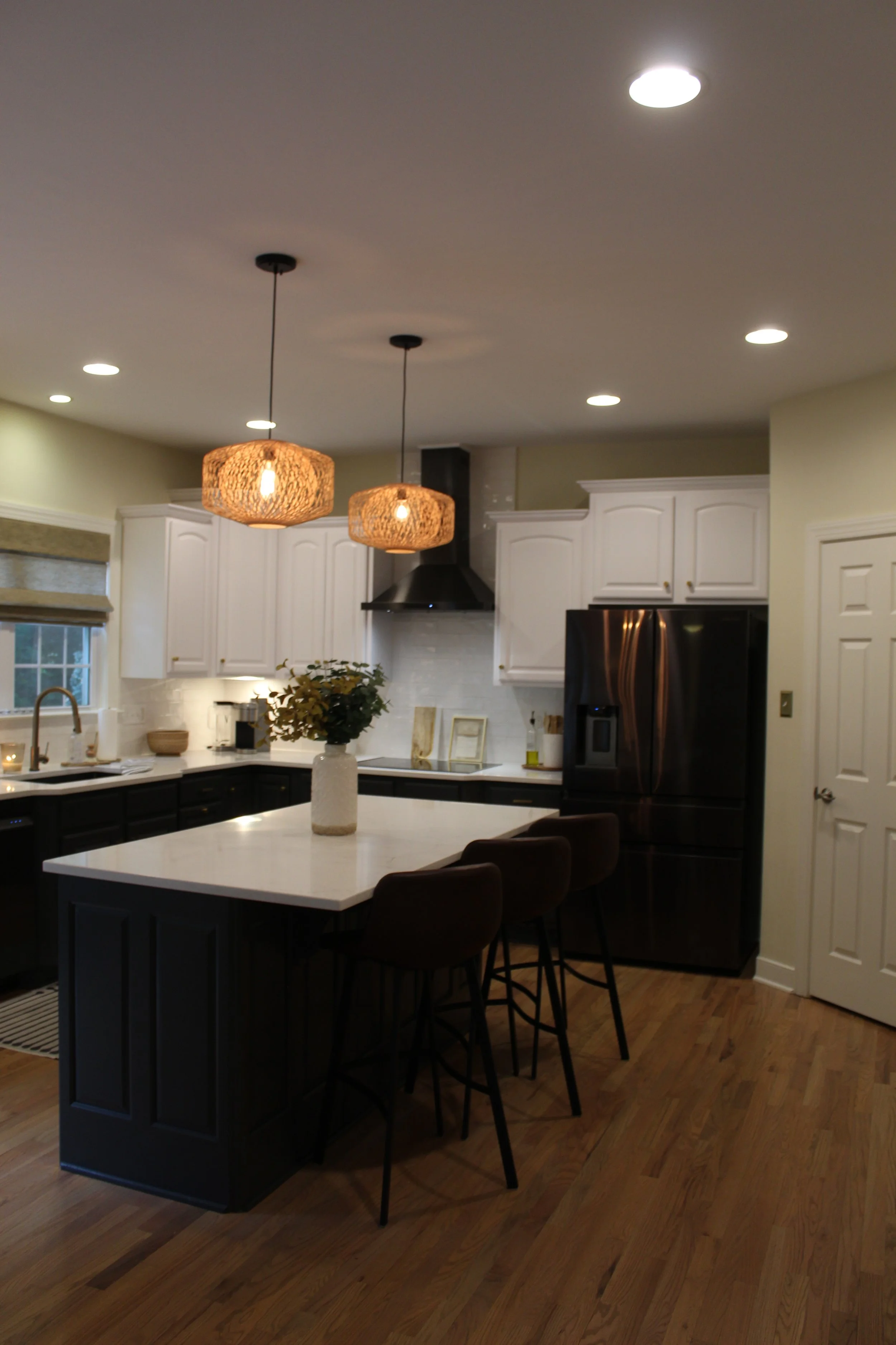 Modern kitchen with white upper cabinets, dark lower cabinets, a kitchen island with a white countertop, three brown barstools, black refrigerator, black range hood, and two decorative pendant lights.