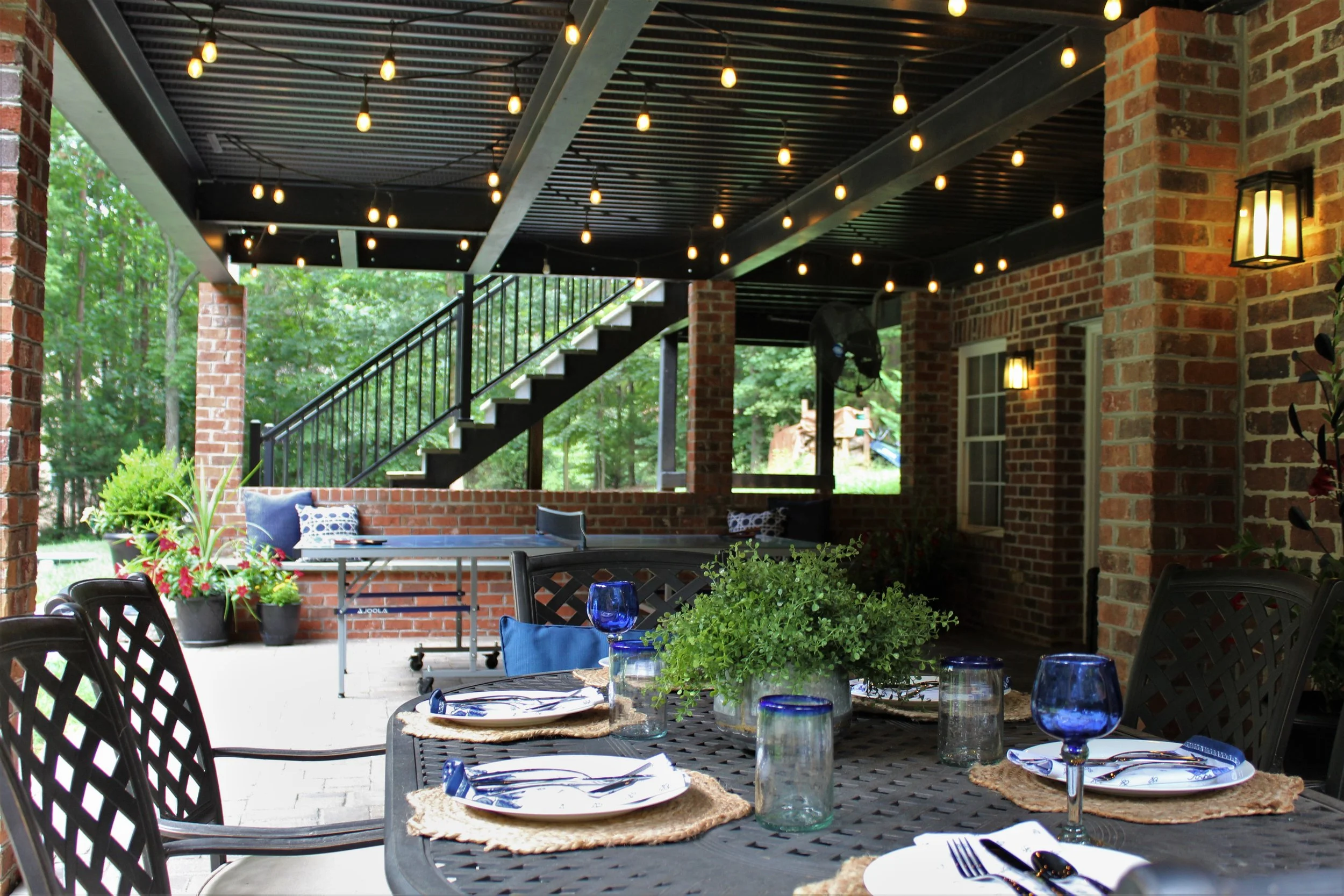 Covered outdoor patio with a black metal dining table set for four, decorated with blue glassware, white plates, and greenery. In the background, there is a brick wall, outdoor stairs, string lights overhead, and lush green trees surrounding the spac