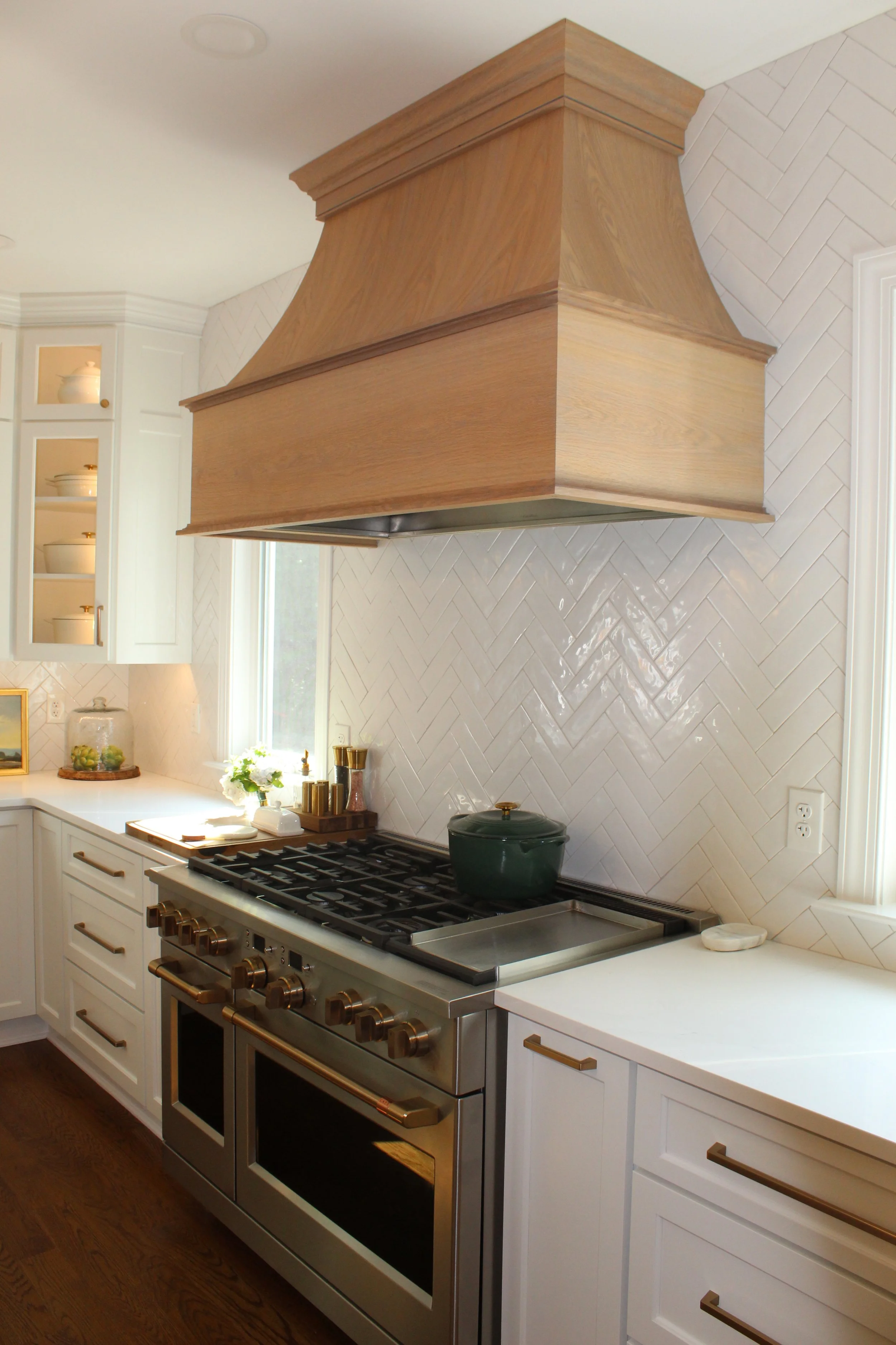 A modern kitchen with white cabinets, a large stainless steel stove and oven, and a wooden range hood. There is a window with sunlight, decorative dishes, and kitchen utensils on the counter.