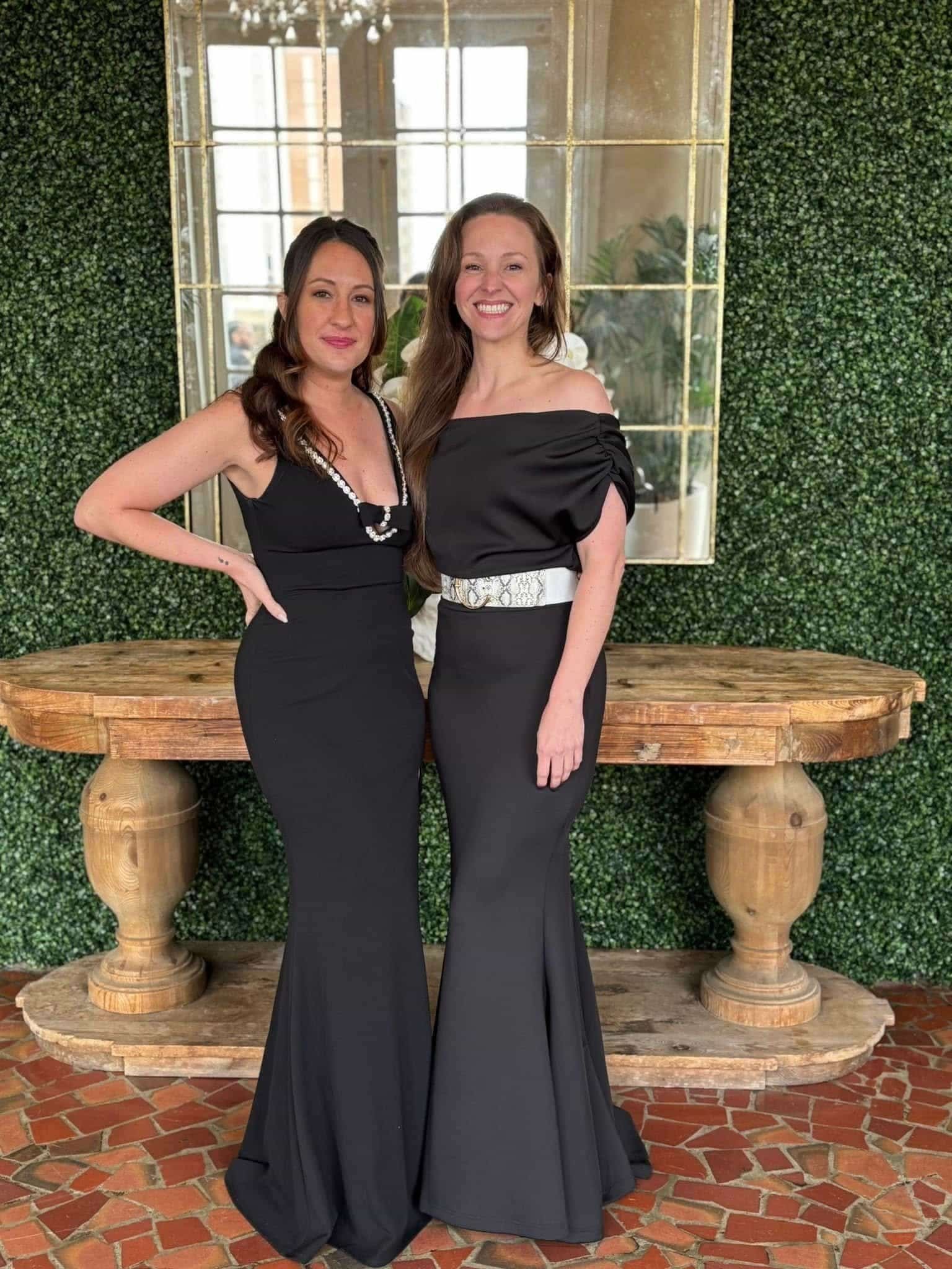 Two women in black dresses standing side by side, smiling, in front of a vintage-style mirror with gold frame and a trimmed green hedge wall background, on a brick floor, with a rustic wooden table below.