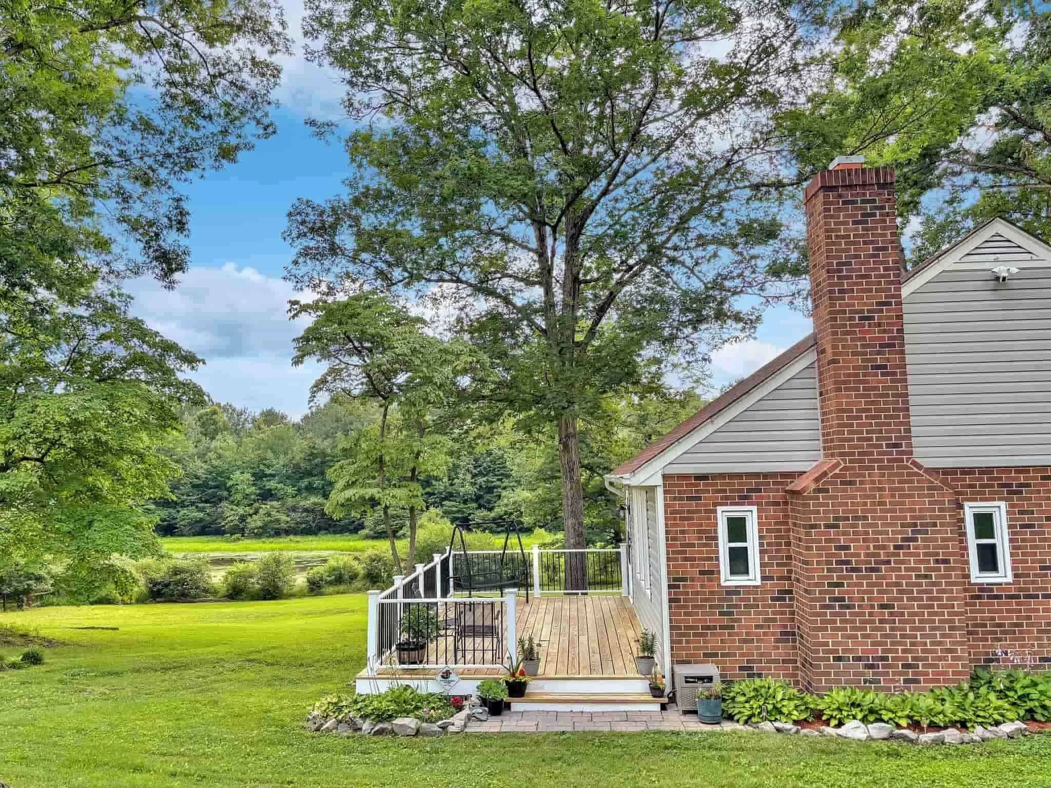 Brick house with a deck overlooks a spacious green lawn, surrounded by trees under a partly cloudy blue sky.