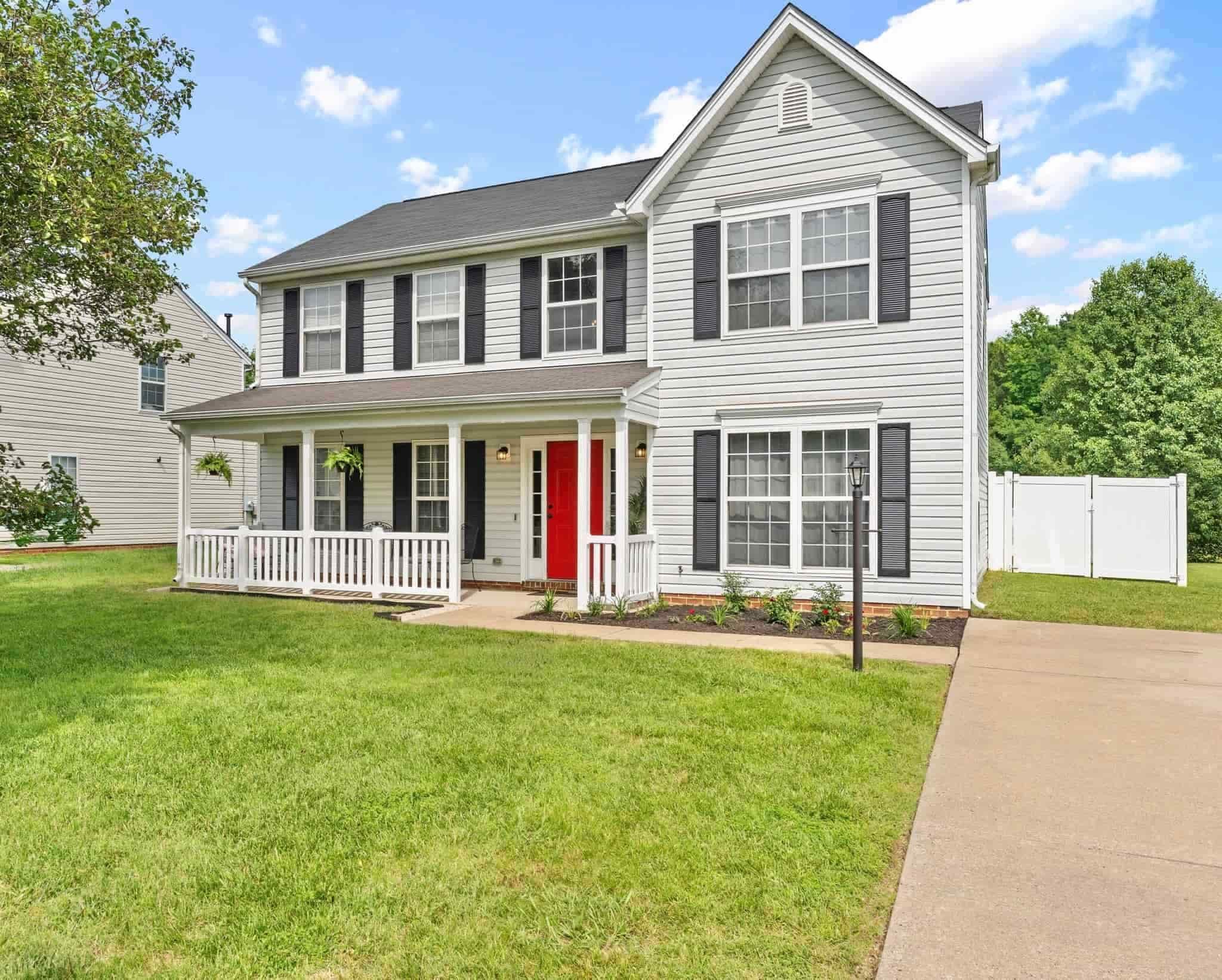 Two-story house with gray siding, black shutters, a bright red door, white porch, and a driveway, set on a green lawn.