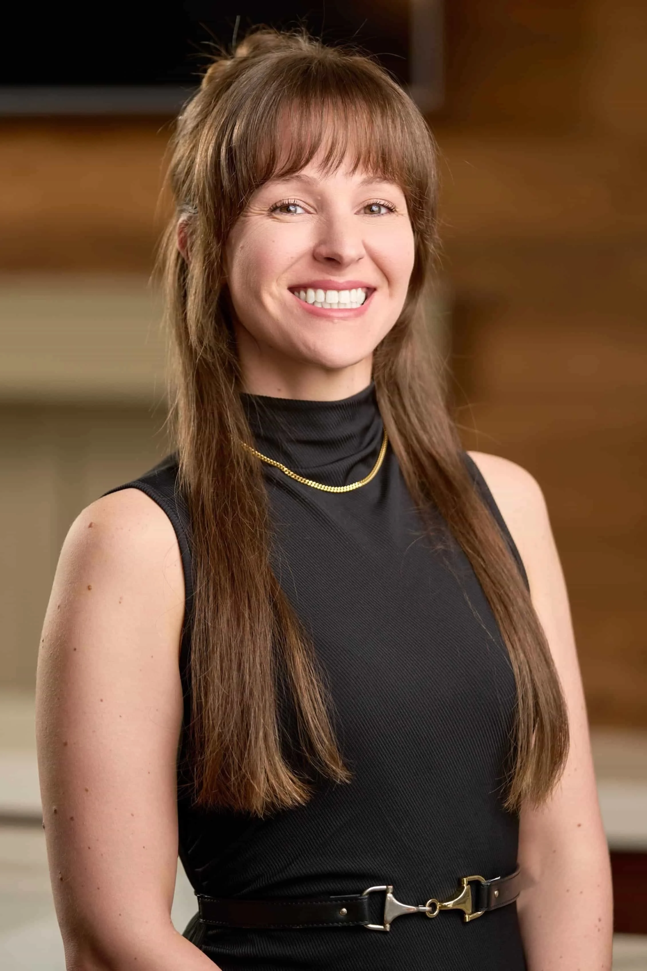 A woman with long brown hair, smiling, wearing a black sleeveless top, a gold necklace, and a black belt with a gold buckle, standing indoors with a wooden background.