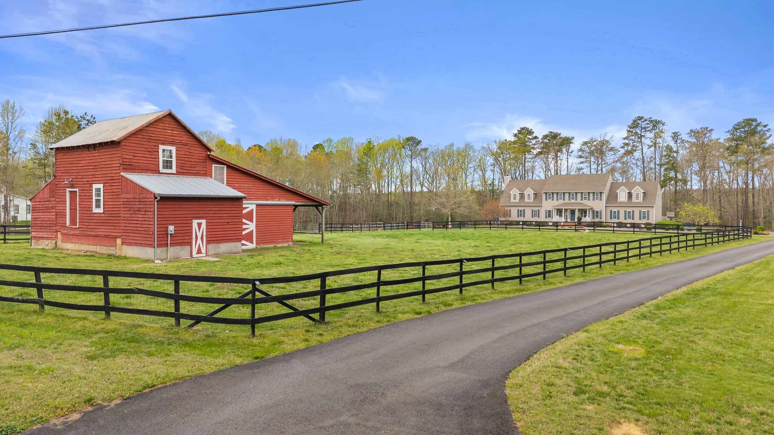 A red barn with a metal roof and a modern house in the background, separated by a black wooden fence and a paved driveway, with green grass and trees under a blue sky.