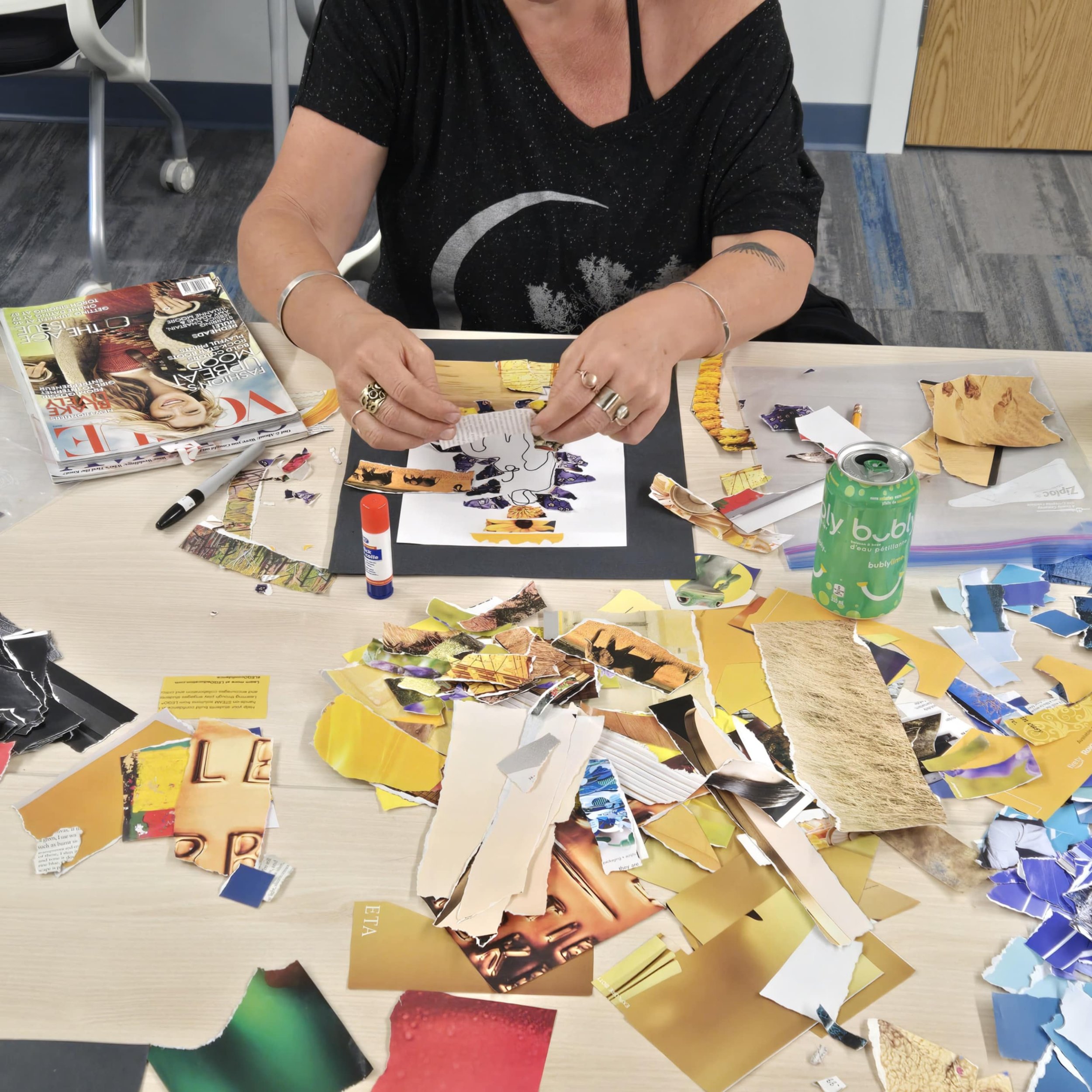 Woman creating art with torn magazine pieces at a table covered in colorful paper scraps, surrounded by a glue stick, marker, magazine, and soda can.