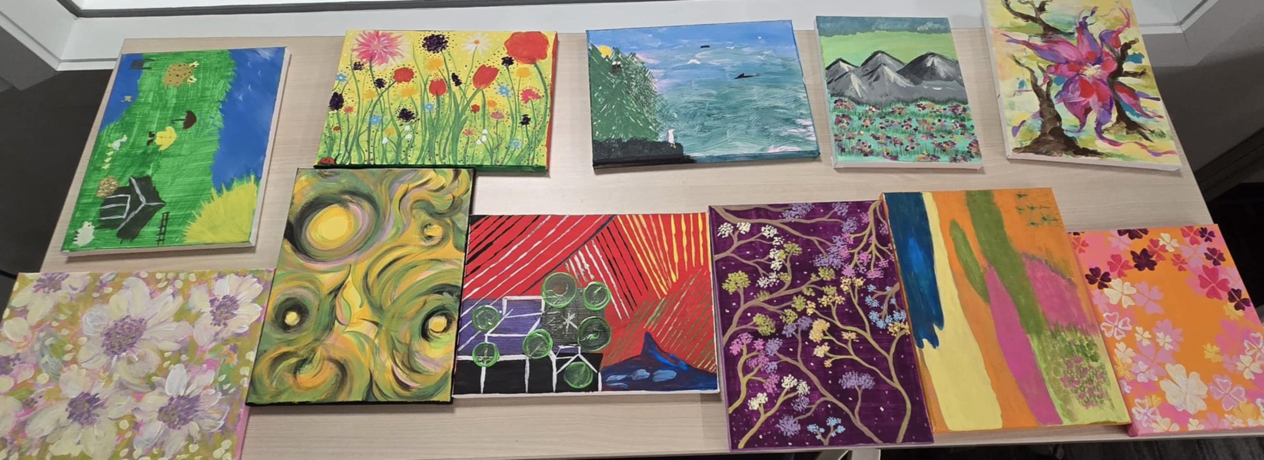 A collection of colorful paintings spread out on a table, depicting various nature scenes and abstract designs.
