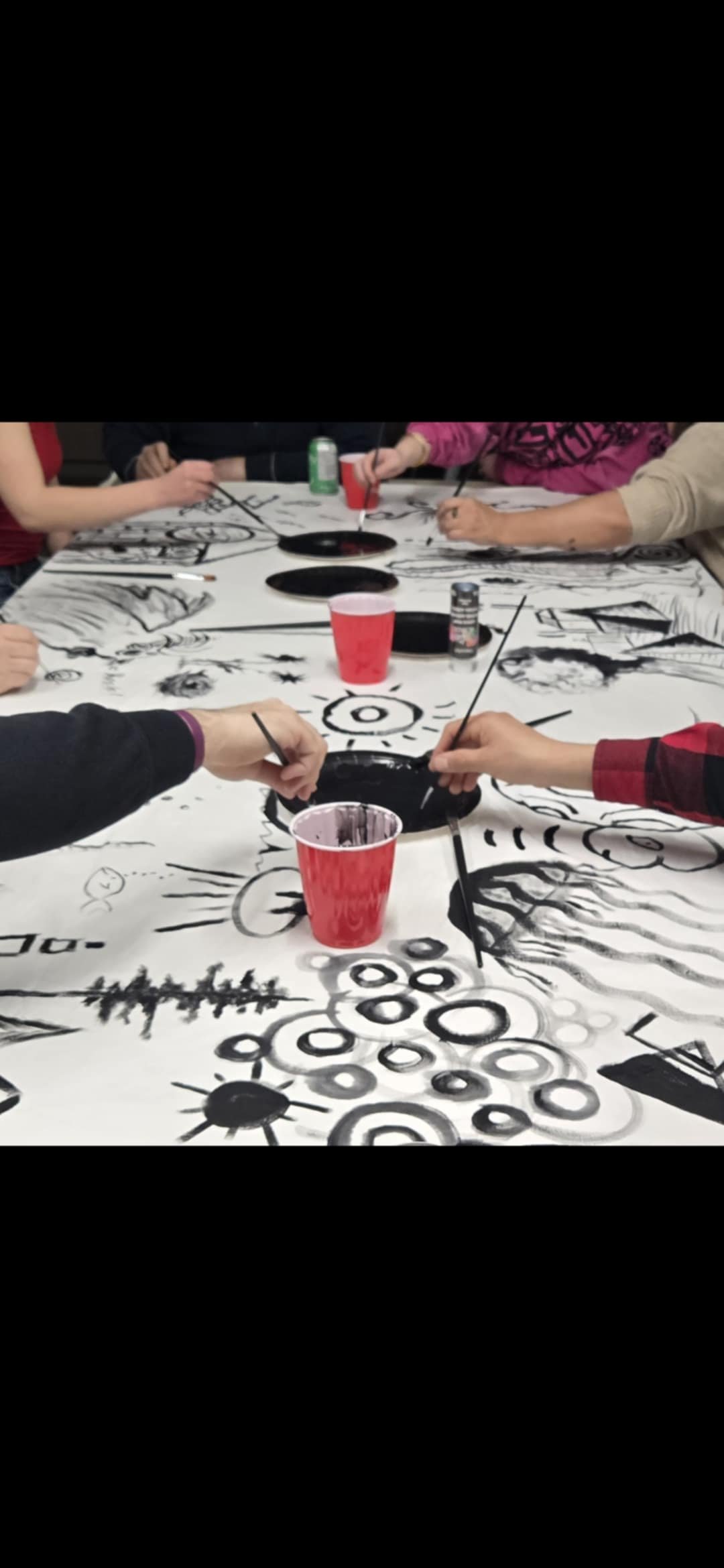 Group of people painting a large, abstract black and white mural on a long table with cups of paint.