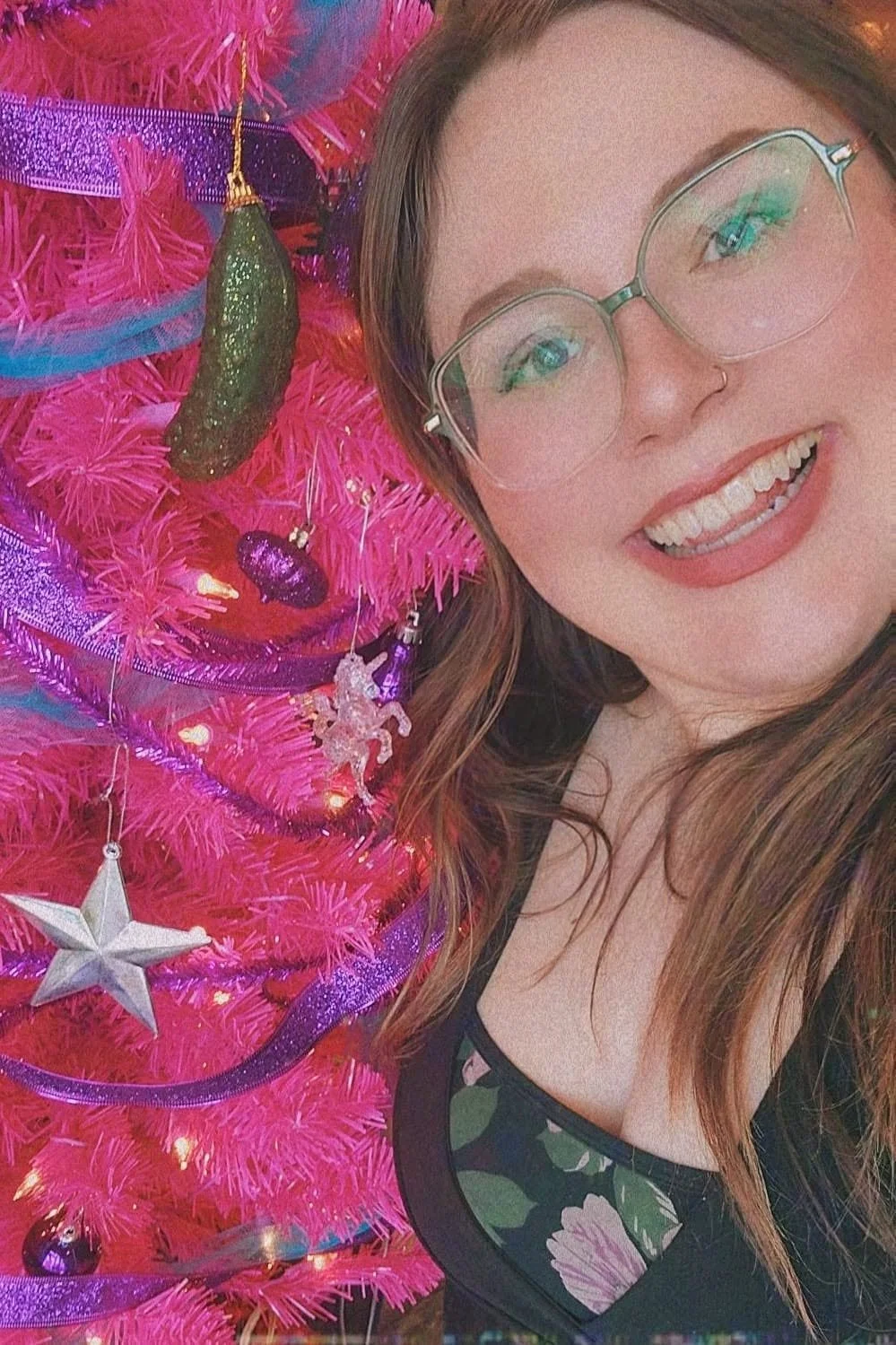 A woman with glasses and a nose ring taking a selfie next to a pink Christmas tree decorated with various ornaments and ribbons.